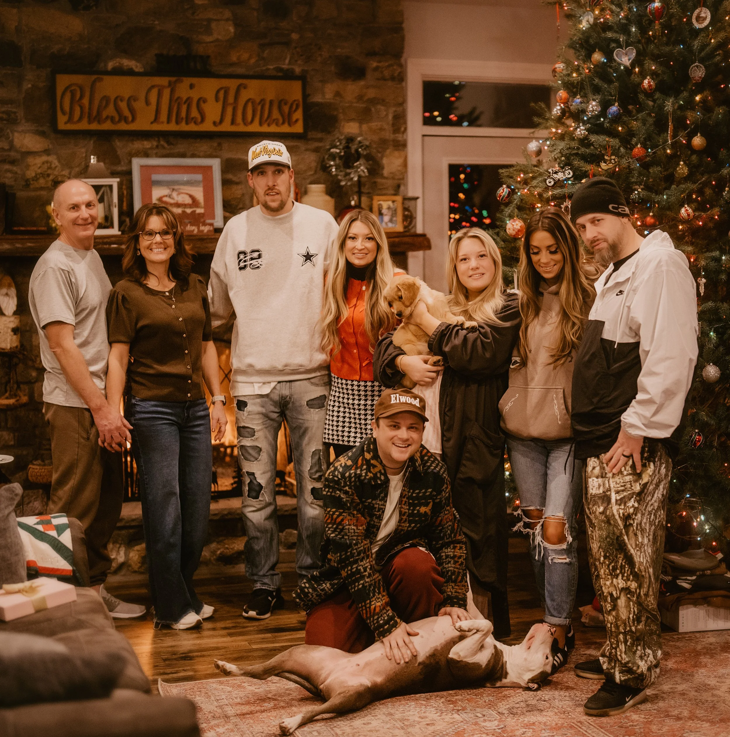 A group of nine people and a dog are gathered in a cozy living room decorated for Christmas, standing in front of a fireplace and a large Christmas tree with ornaments and lights. They are smiling and posing for the photo. There is a sign that says 'Bless This House' above the fireplace.