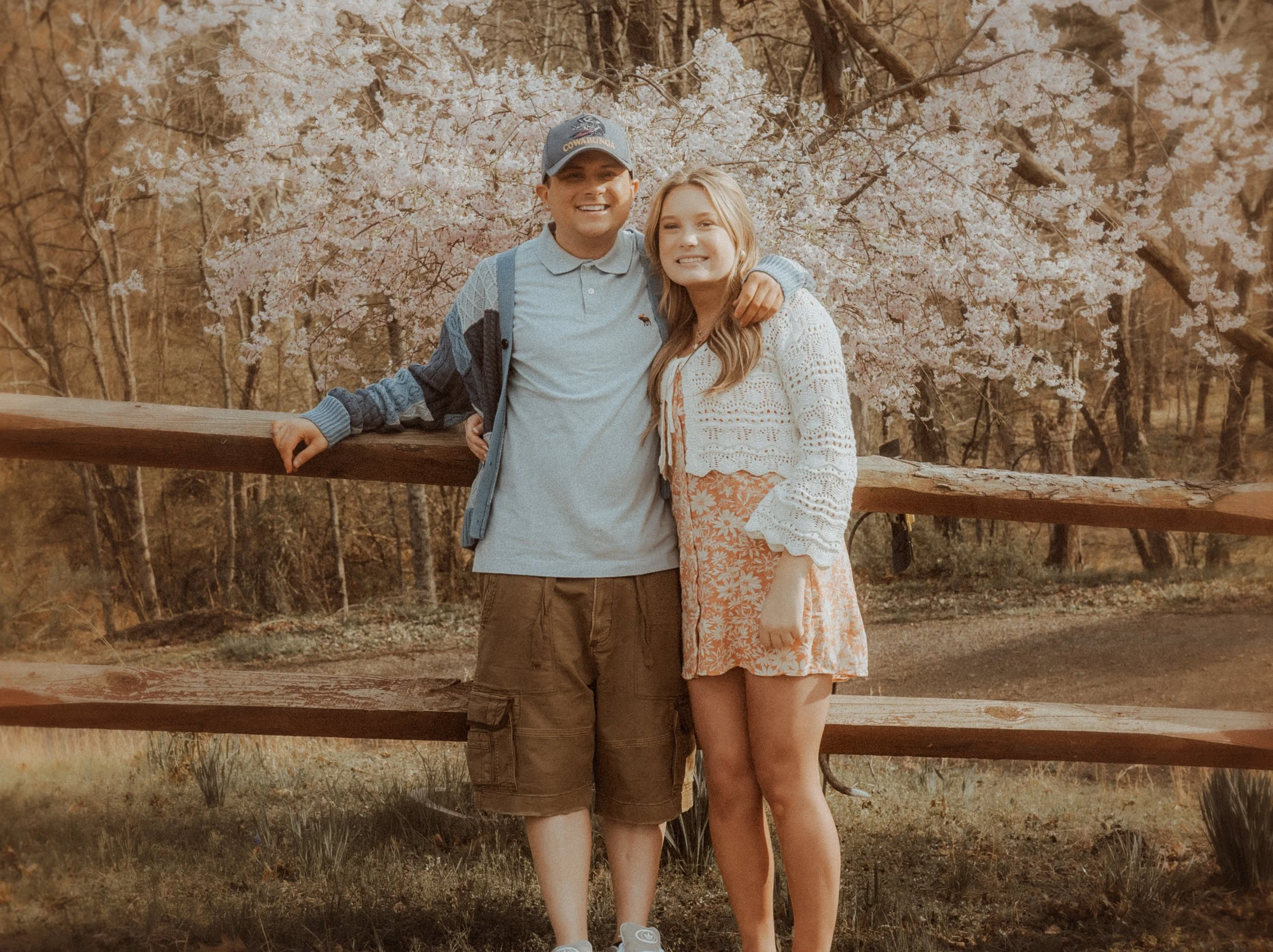 A young man and woman smiling together outdoors near a blooming cherry blossom tree, standing in front of a wooden fence during spring.