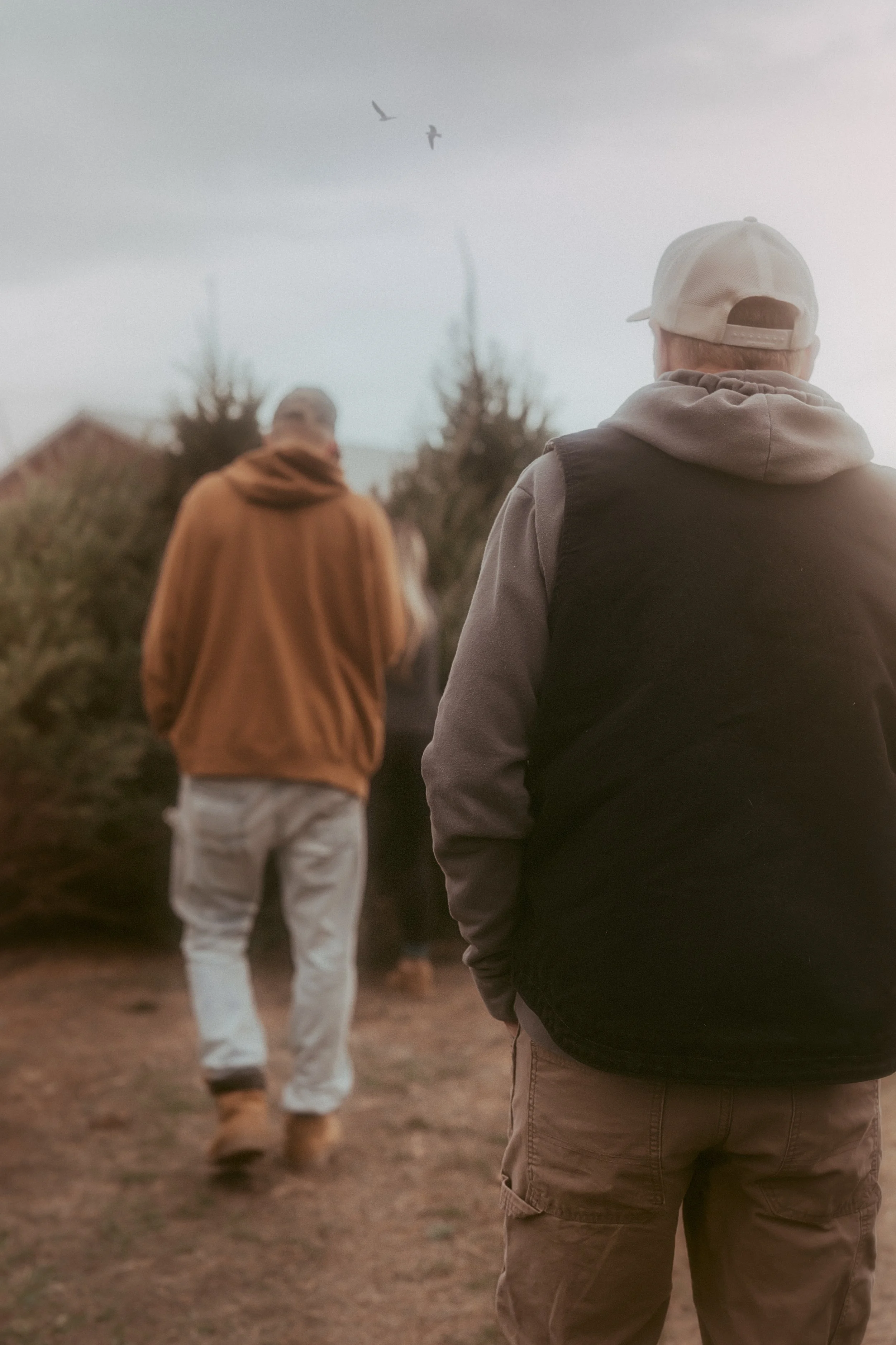 Three men walking outdoors during daytime with trees and houses in the background, one is in the foreground wearing a beige cap and grey hoodie, the other two in the background are wearing a brown jacket and a black hoodie.
