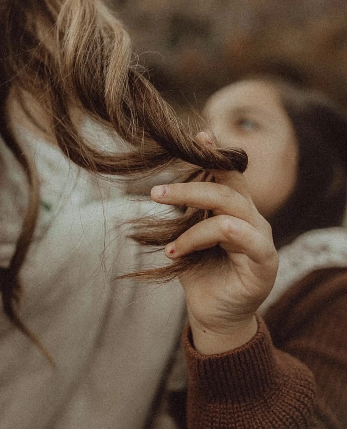 A close-up of a person's hand with slightly reddened fingertips, holding a lock of curly hair. The person appears to be lying down, with their face blurred in the background.
