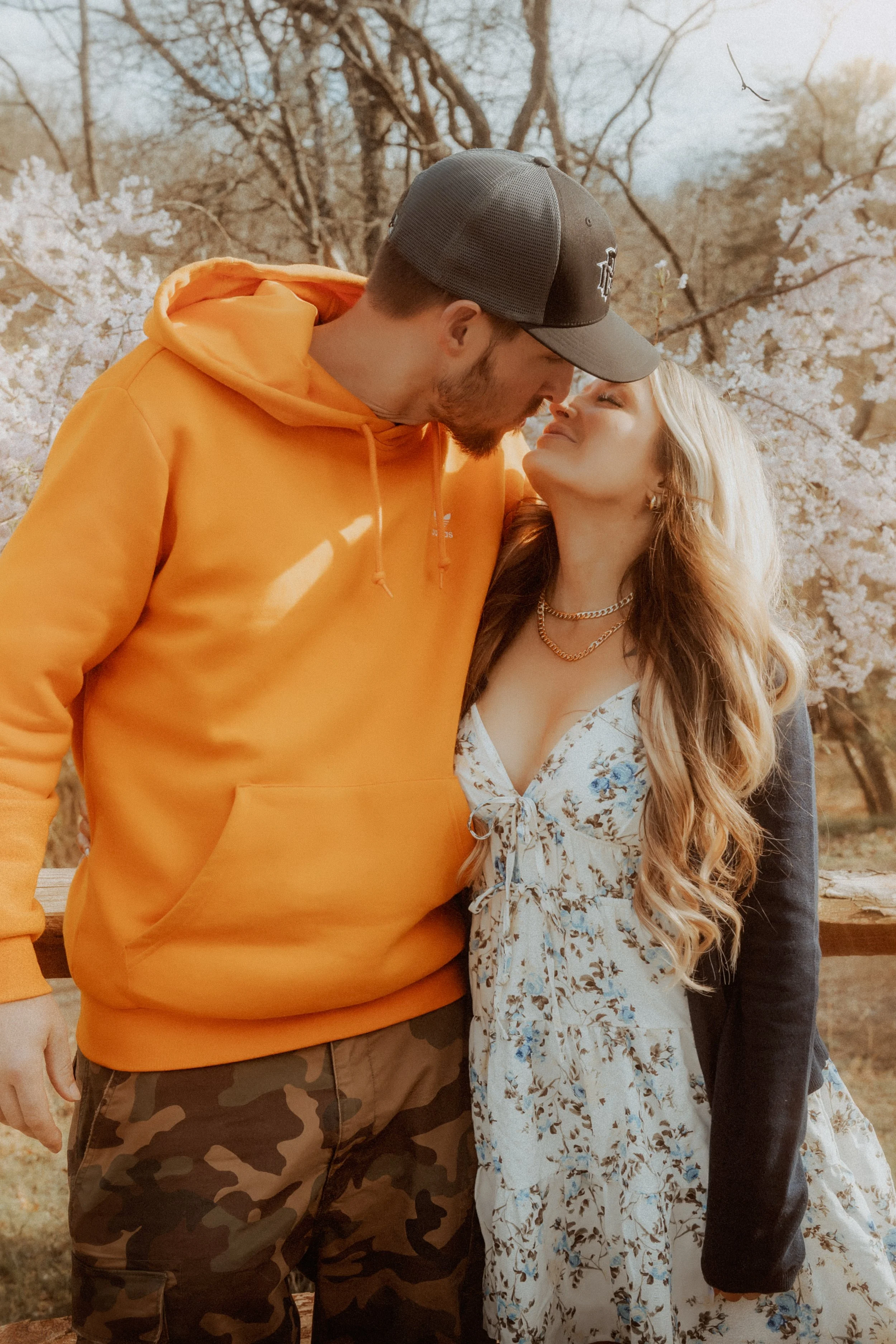 A young man in an orange hoodie and camouflage pants leaning close to a young woman in a floral dress and a black jacket, with their foreheads touching. They are outdoors among blooming trees in spring.