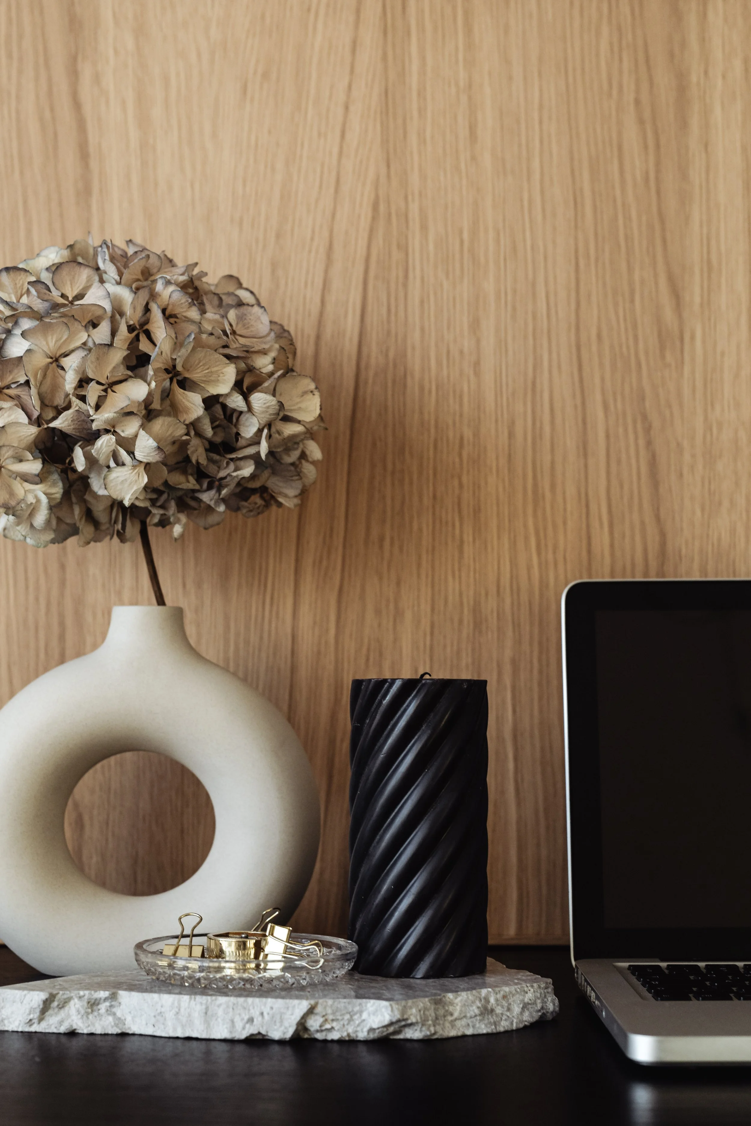 Decorative home office desk with a dried hydrangea flower in a white vase, a black twisted candle, gold paper clips in a small glass dish, and a laptop on a marble tray against a wooden wall.