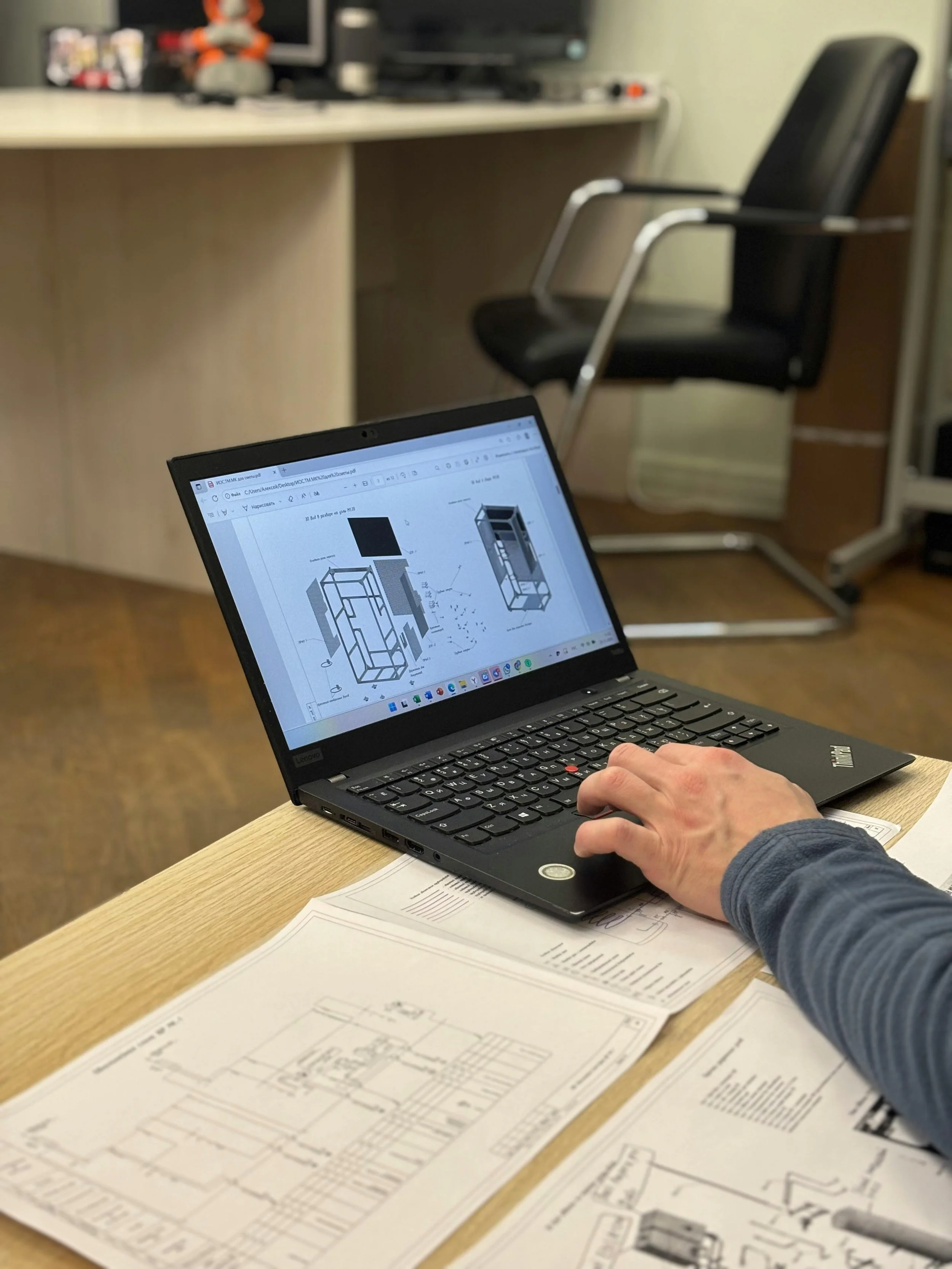 A person working on architectural blueprints with a laptop displaying 3D building models. The workspace includes a desk with papers and architectural drawings, a black office chair, and a wall with a monitor and decorative items in the background.