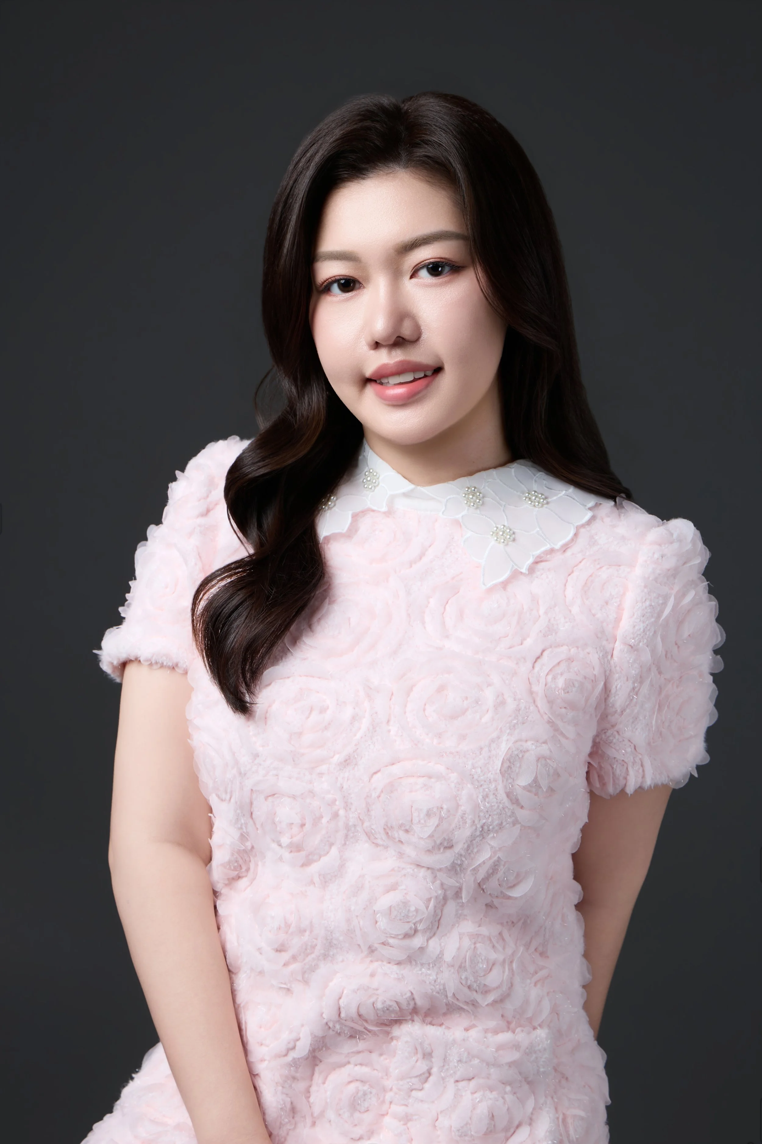 A woman with long dark hair styled in loose waves, wearing a light pink textured dress with a white lace collar adorned with small pearls, smiling gently against a dark gray background.