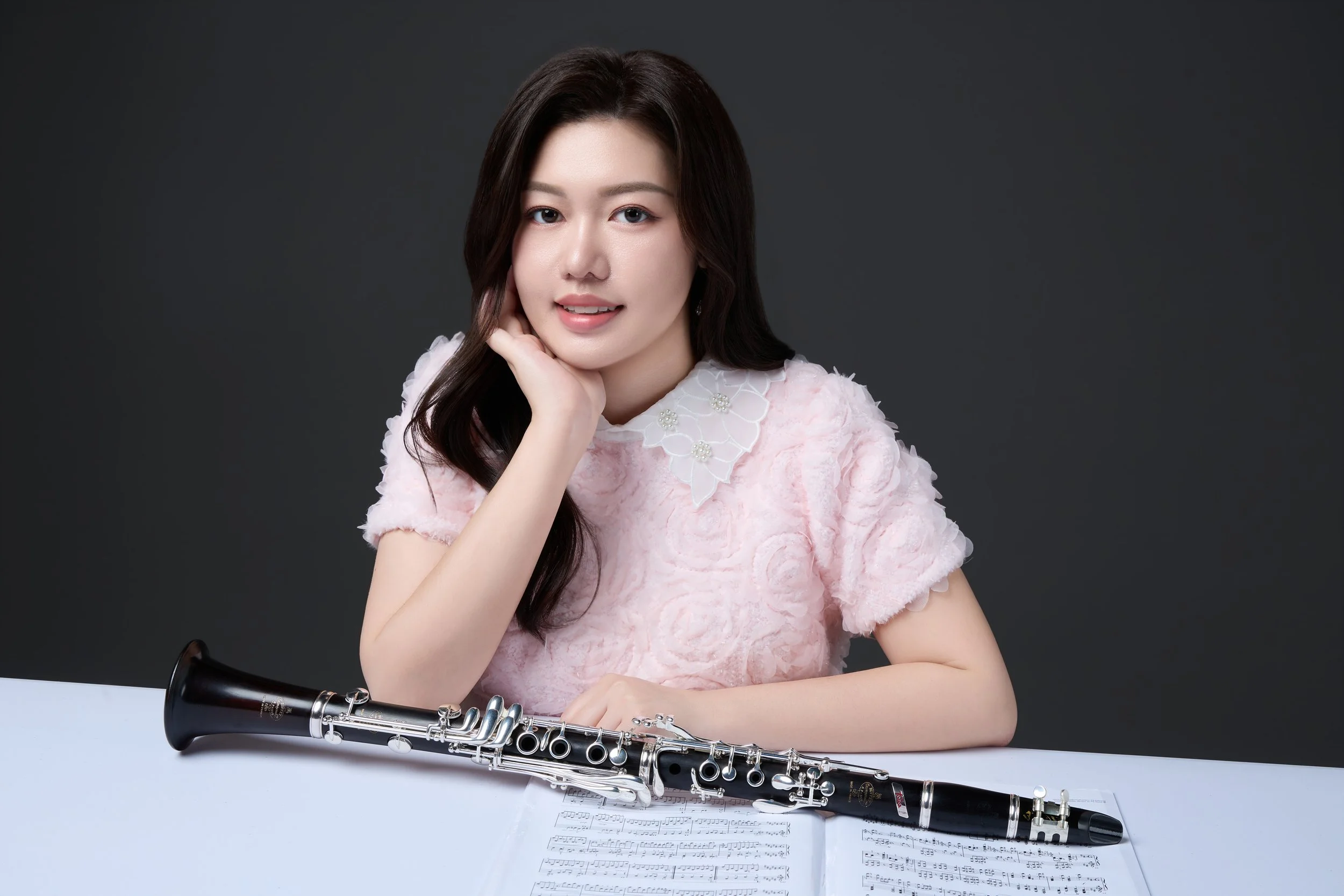 A woman with dark hair, wearing a pink dress with white lace collar, sitting at a white table with a clarinet and sheet music, posing against a dark gray background.