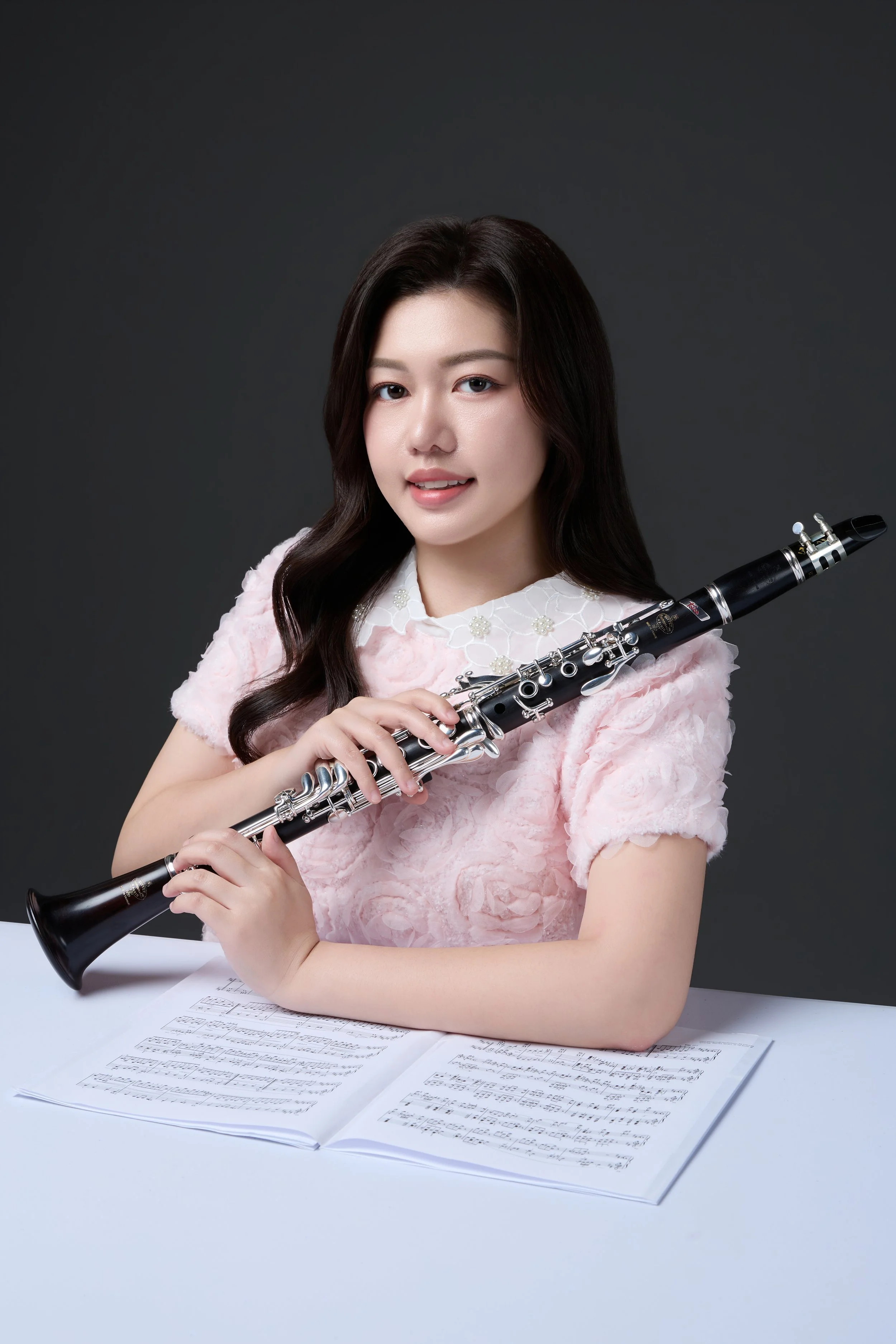 A young woman with long dark hair and fair skin holding a black clarinet, sitting at a white table with sheet music open in front of her, against a dark plain background.