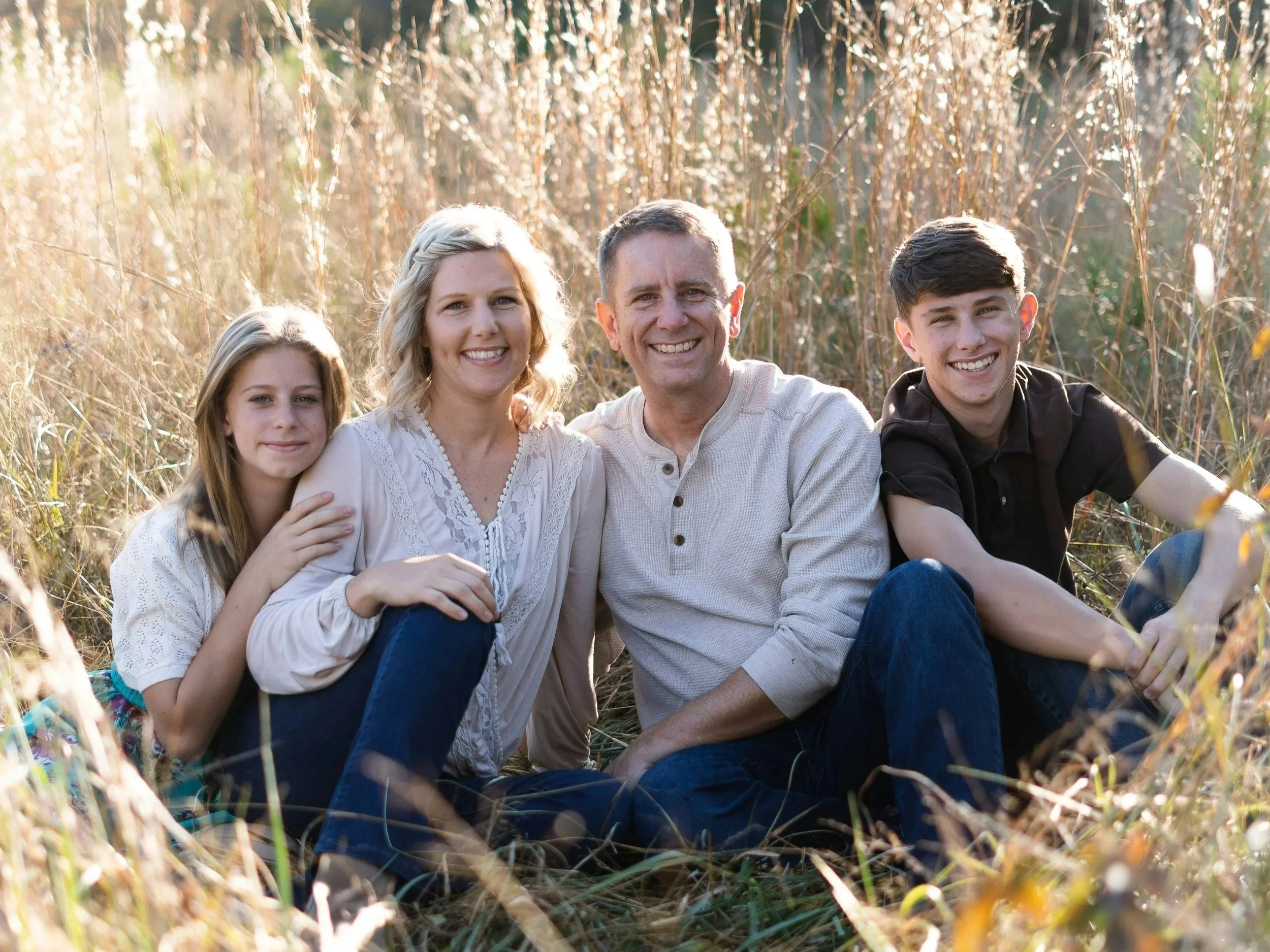 A family of five sitting together outdoors in a field of tall, dry grass on a sunny day. The family includes a woman, a man, two teenage boys, and a young girl. They are smiling and appear happy, enjoying each other's company.