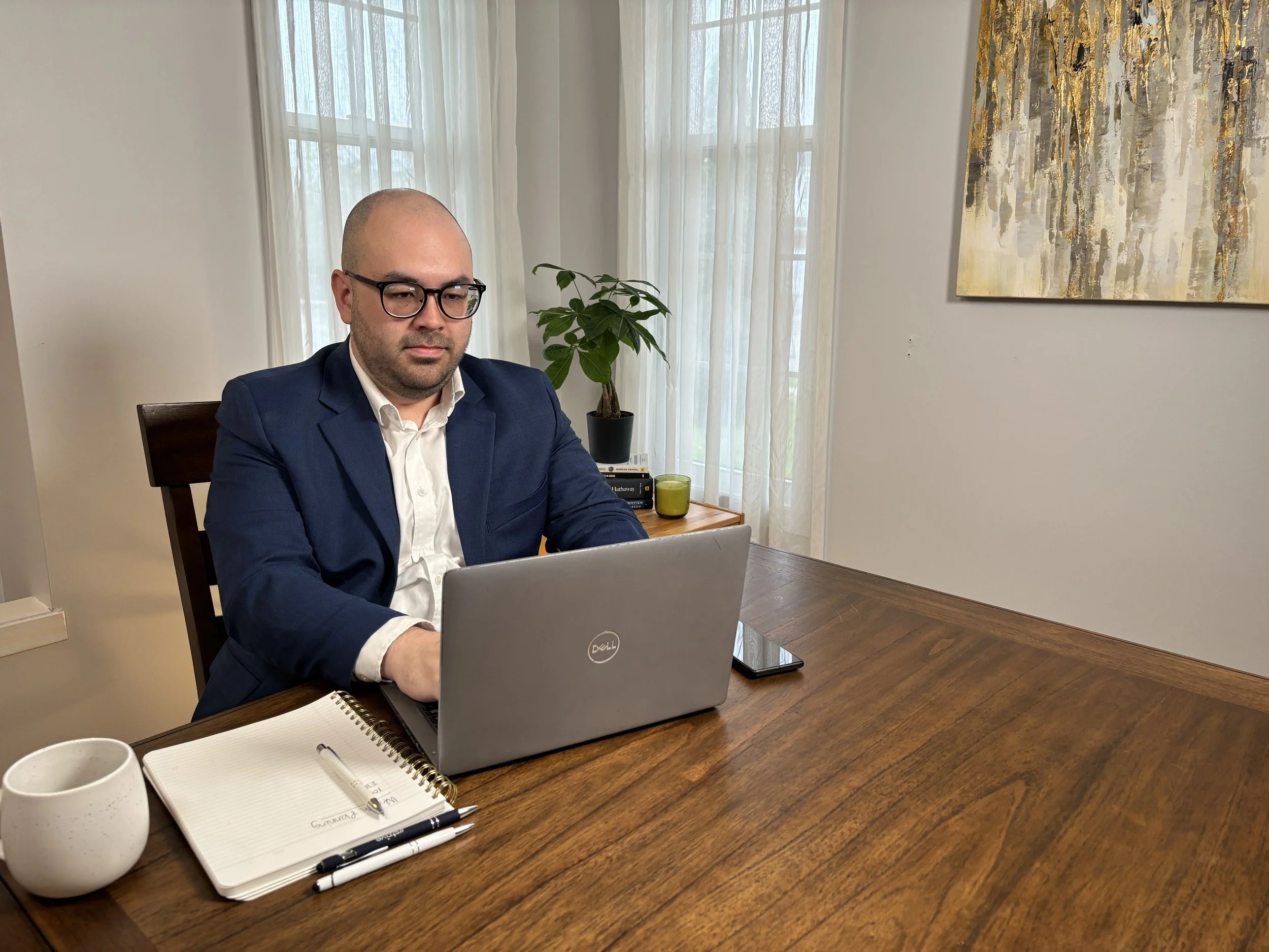 A man in a dark suit and glasses sitting at a wooden table with a laptop, notebooks, pens, a yellow mug, and a white speckled mug, in a bright room with large windows, sheer curtains, a houseplant, and abstract art on the wall.