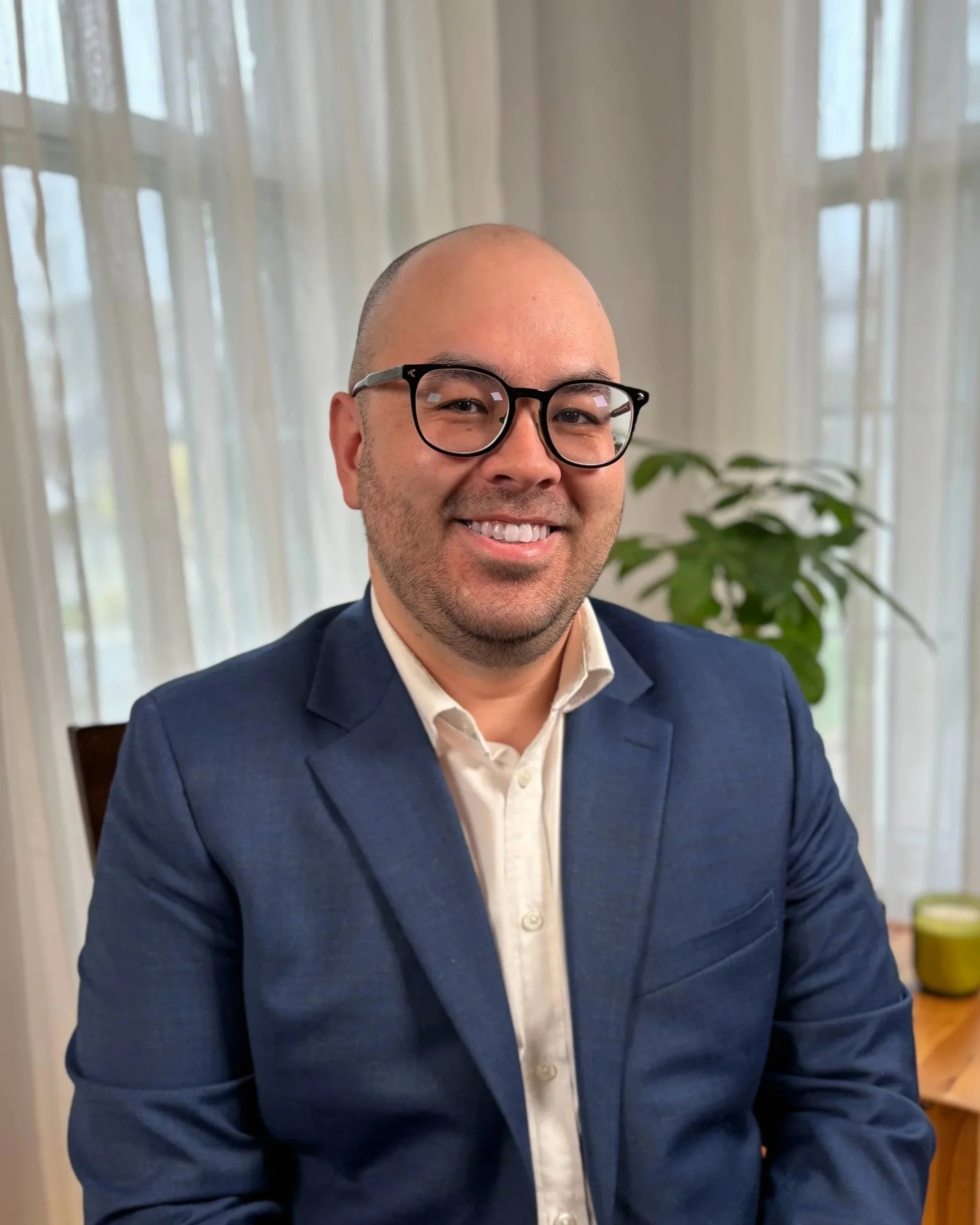 A smiling man with glasses, wearing a blue suit and white shirt, sitting indoors with curtains and a plant visible in the background.
