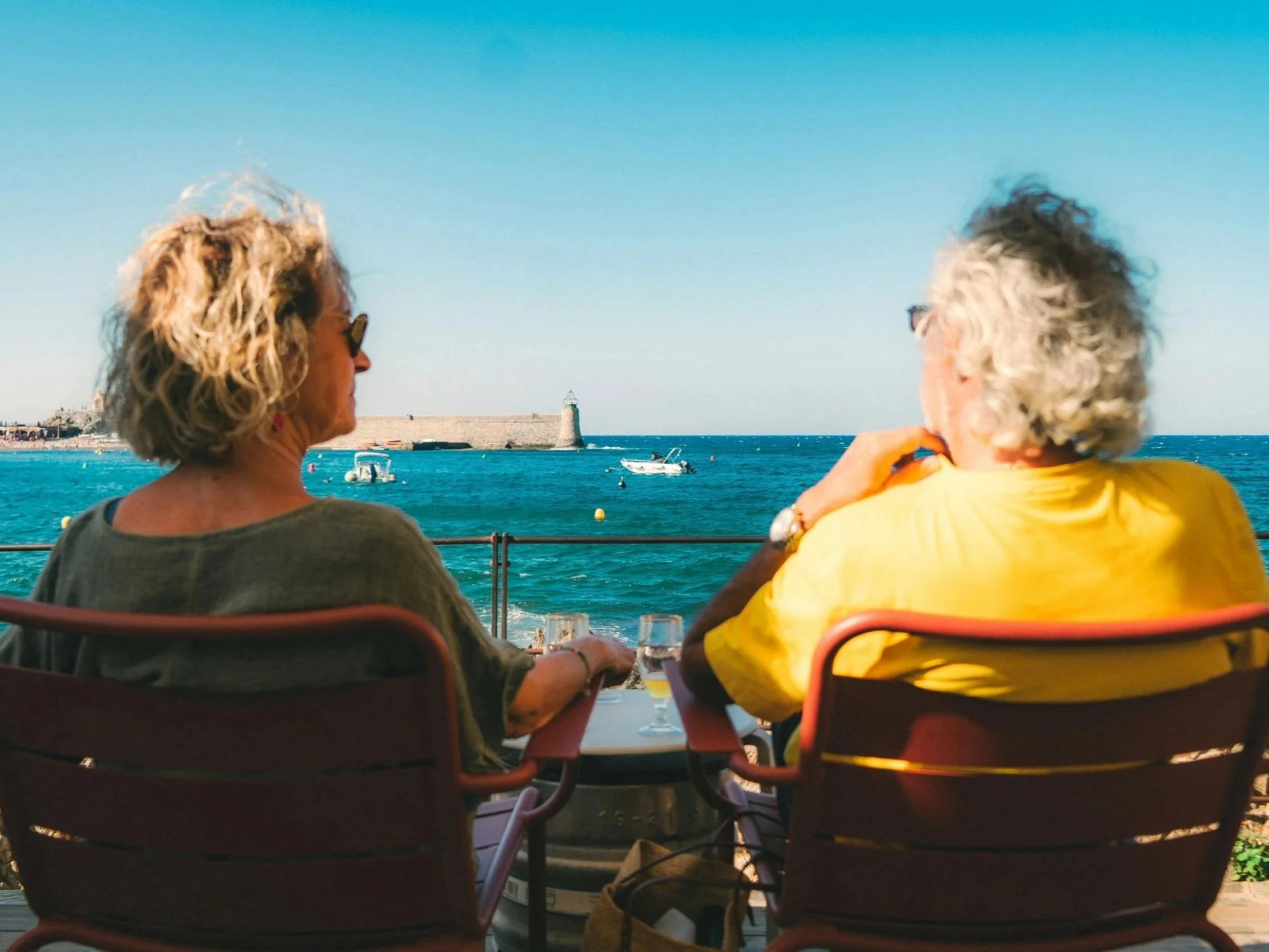 A couple with curly hair sitting at a table by the water, with boats and a lighthouse in the background, enjoying a sunny day.