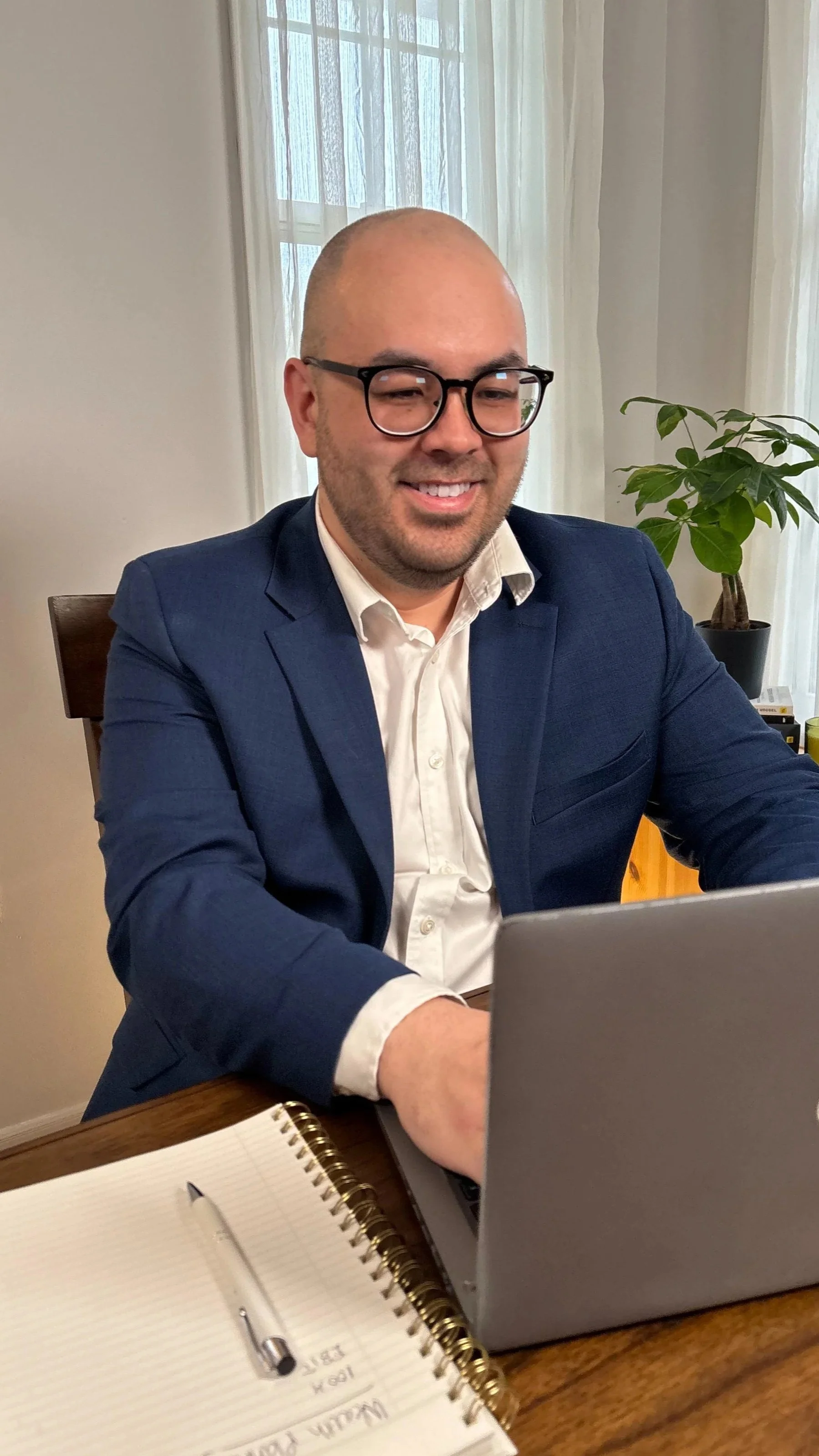 Man in a navy blazer and glasses working on a laptop at a wooden desk, with a notebook and pen. There is a notebook in the foreground and a potted plant in the background.