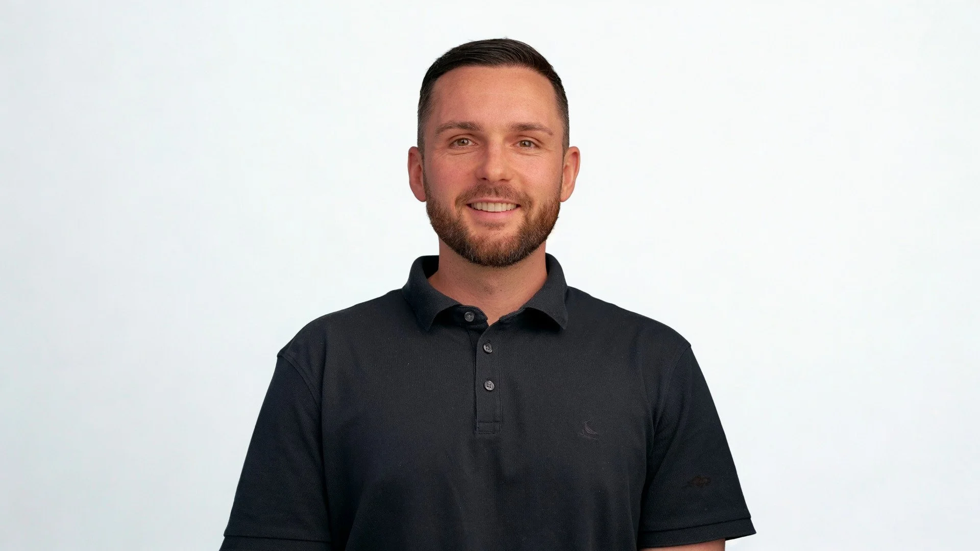 A man with short dark hair and a beard, smiling, wearing a black polo shirt, standing against a plain white background.