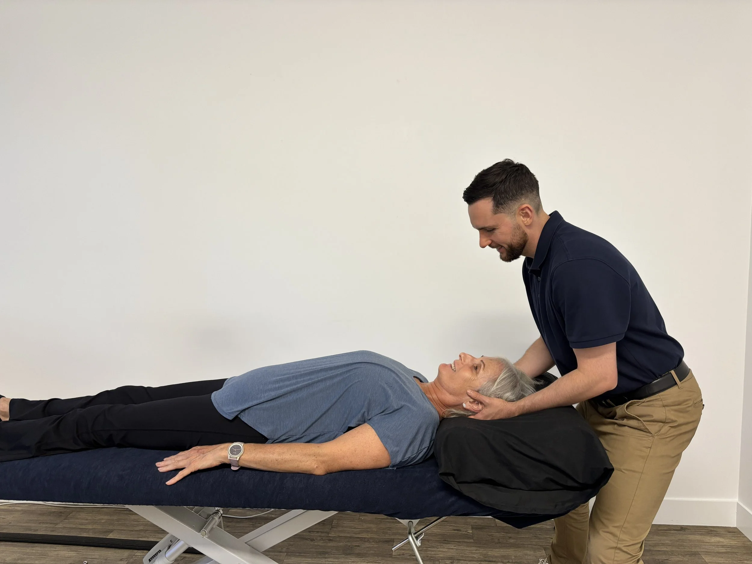 A man with short dark hair and a beard providing CPR to an elderly woman lying on a medical examination table, both smiling, in a room with a plain white wall.
