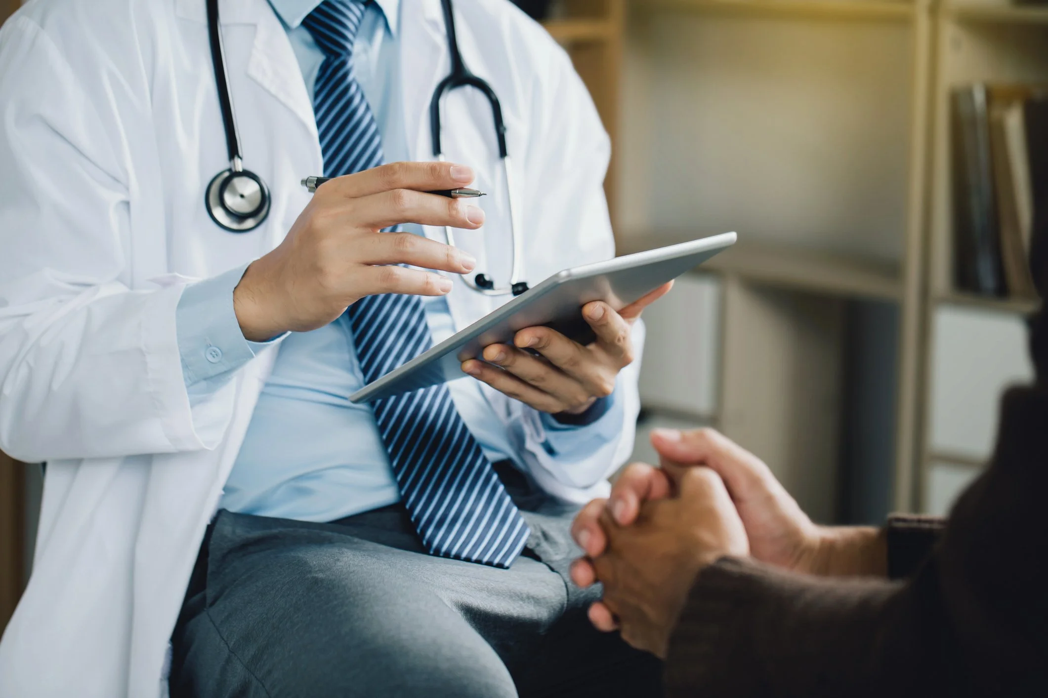 A doctor in a white coat with a stethoscope around his neck is talking to a patient, showing some information on a tablet, in a clinical setting.
