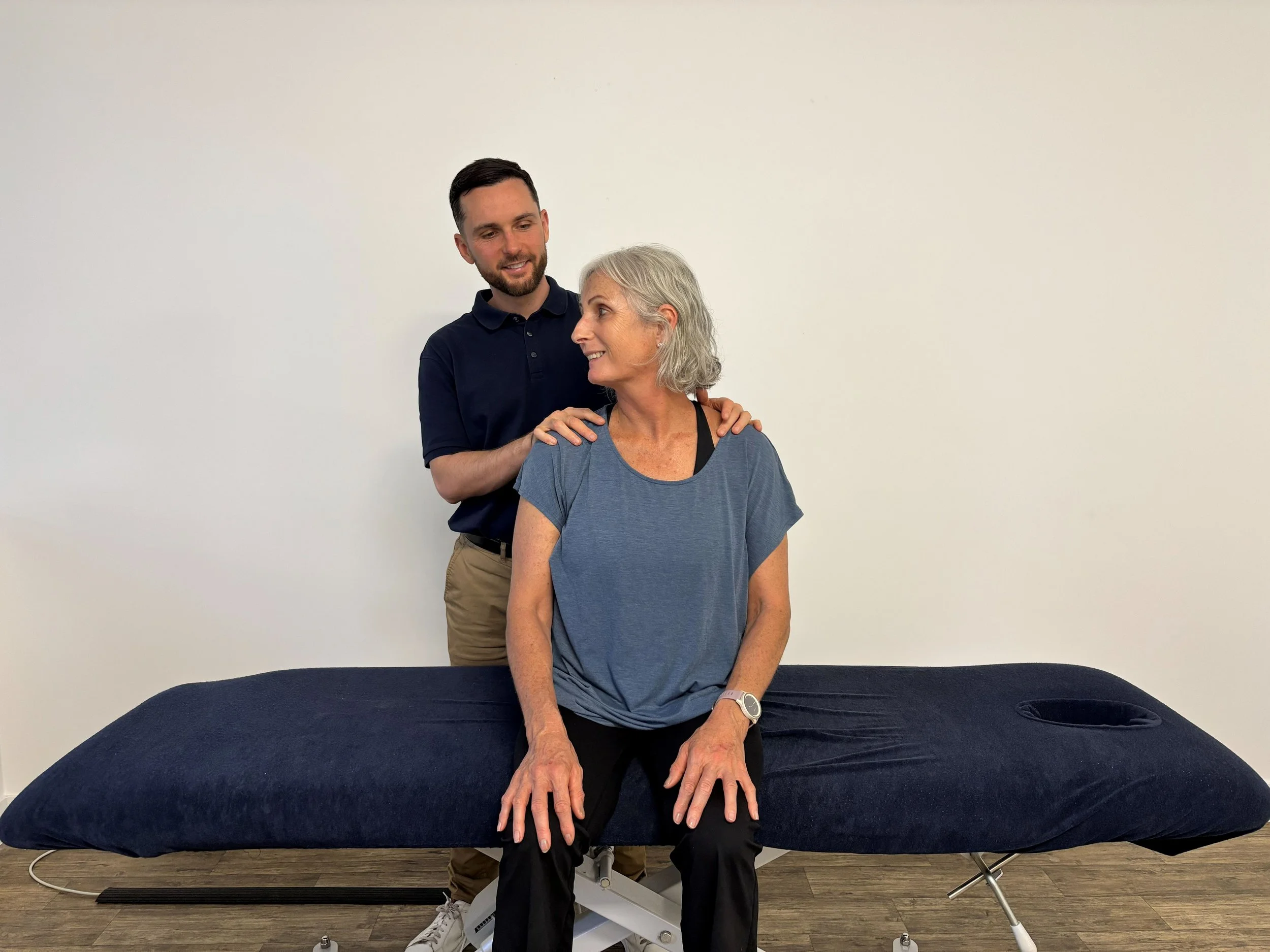 A health care professional, a young man, standing behind an older woman who is seated on an examination table, with his hand on her shoulder, smiling at each other against a plain white wall.