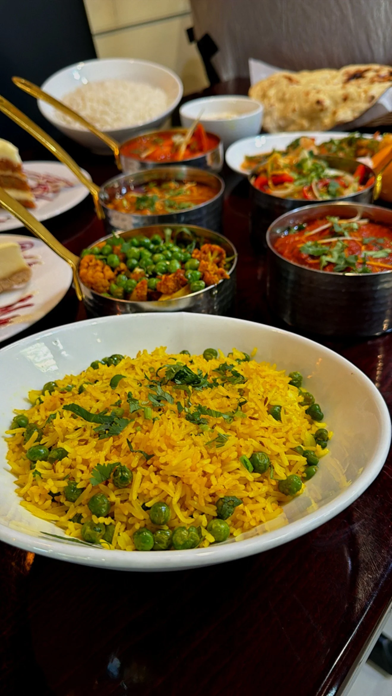 A bowl of yellow rice with green peas garnished with cilantro, surrounded by various Indian dishes and naan bread on a dark wooden table.