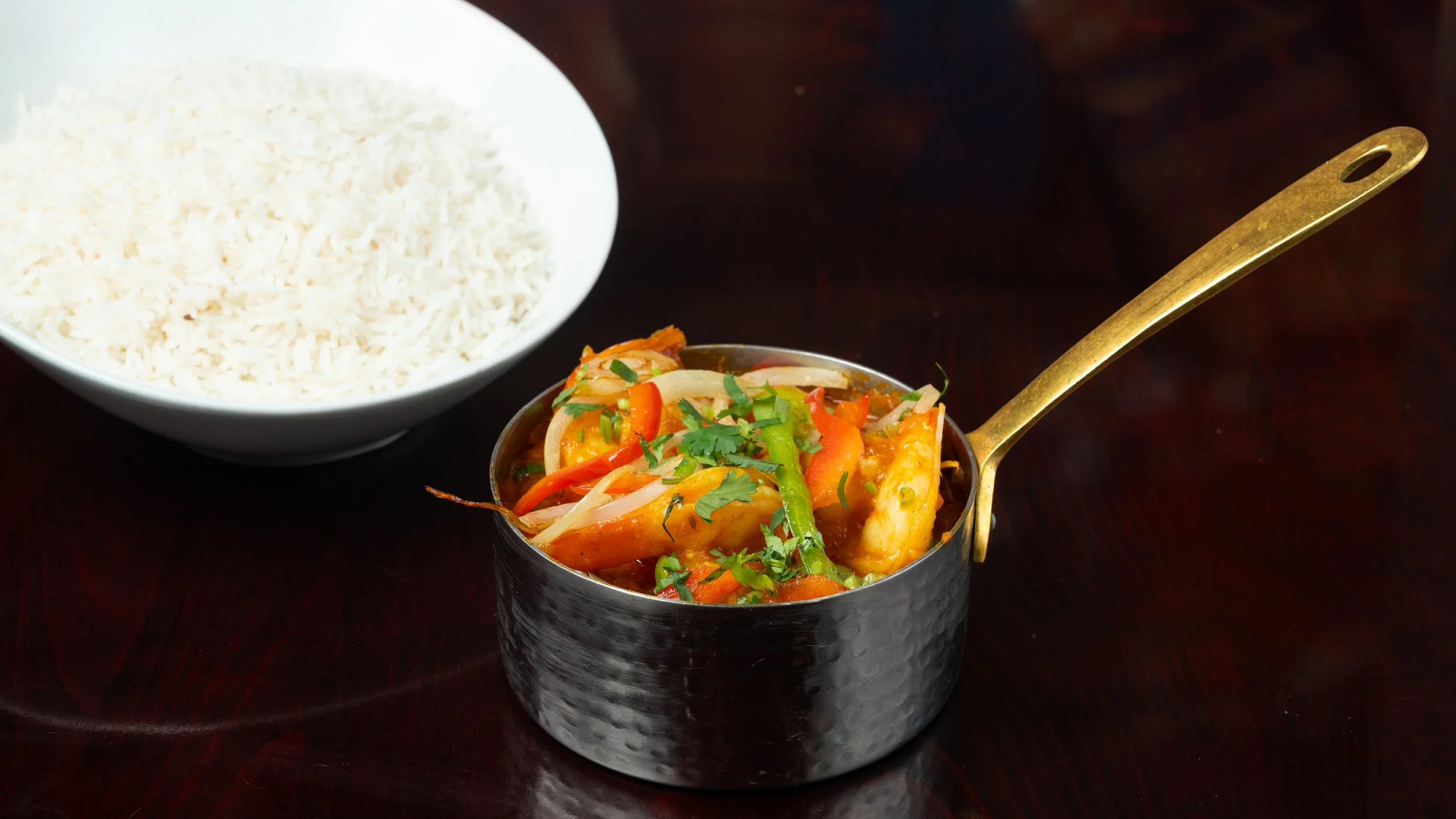 A bowl of white steamed rice and a small pan of vegetable curry with carrots, bell peppers, onions, and cilantro on a dark wooden surface.
