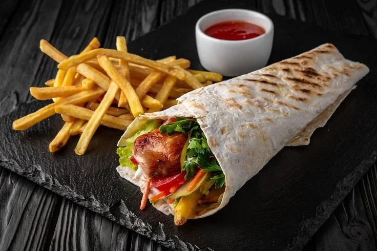 Chicken shawarma wrap with French fries, a side of ketchup, and a small bowl of dipping sauce on a black slate serving board.
