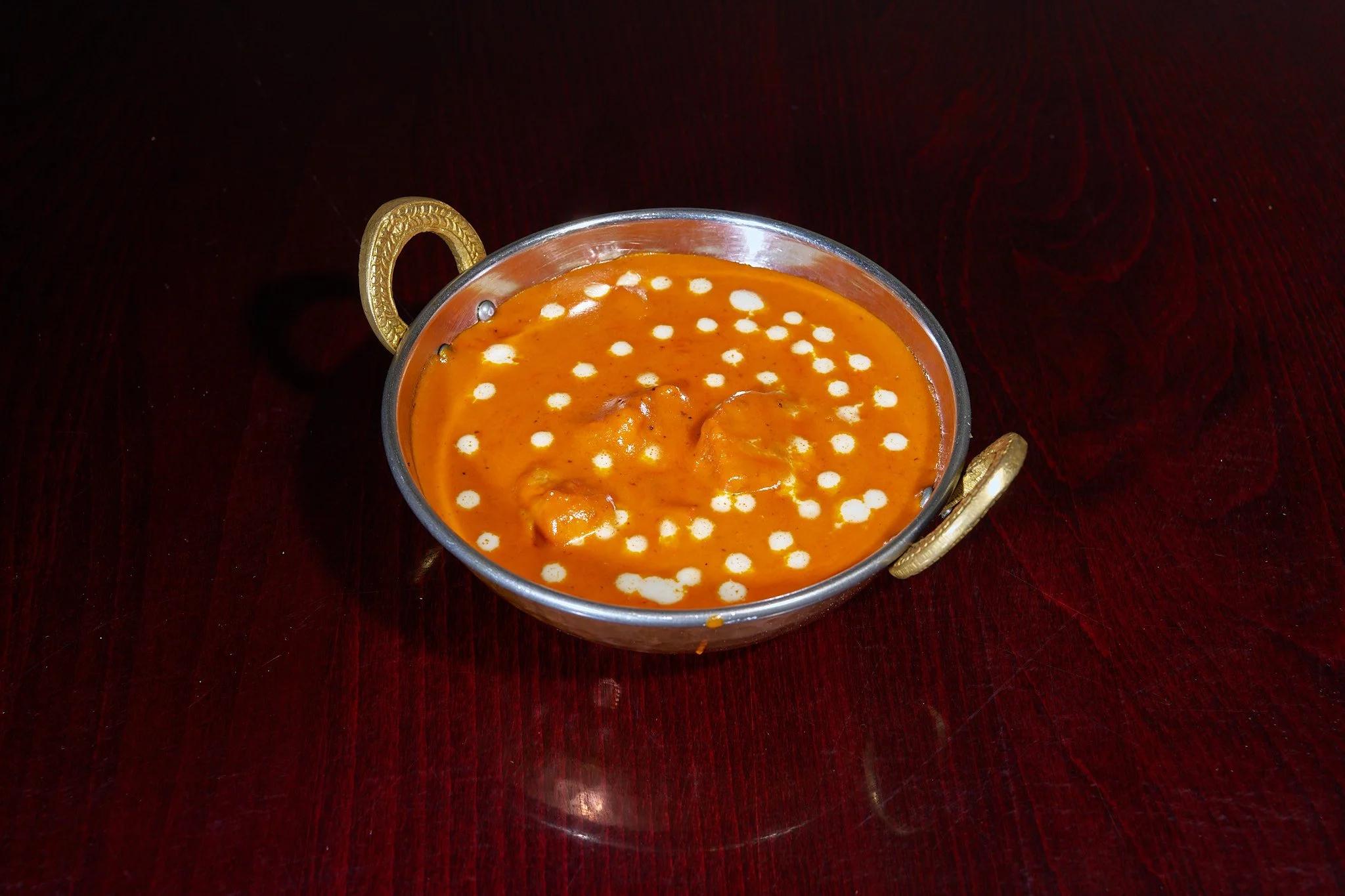 A bowl of Indian butter chicken curry with white cream decor on top, served in a decorative metal bowl on a dark wooden table.