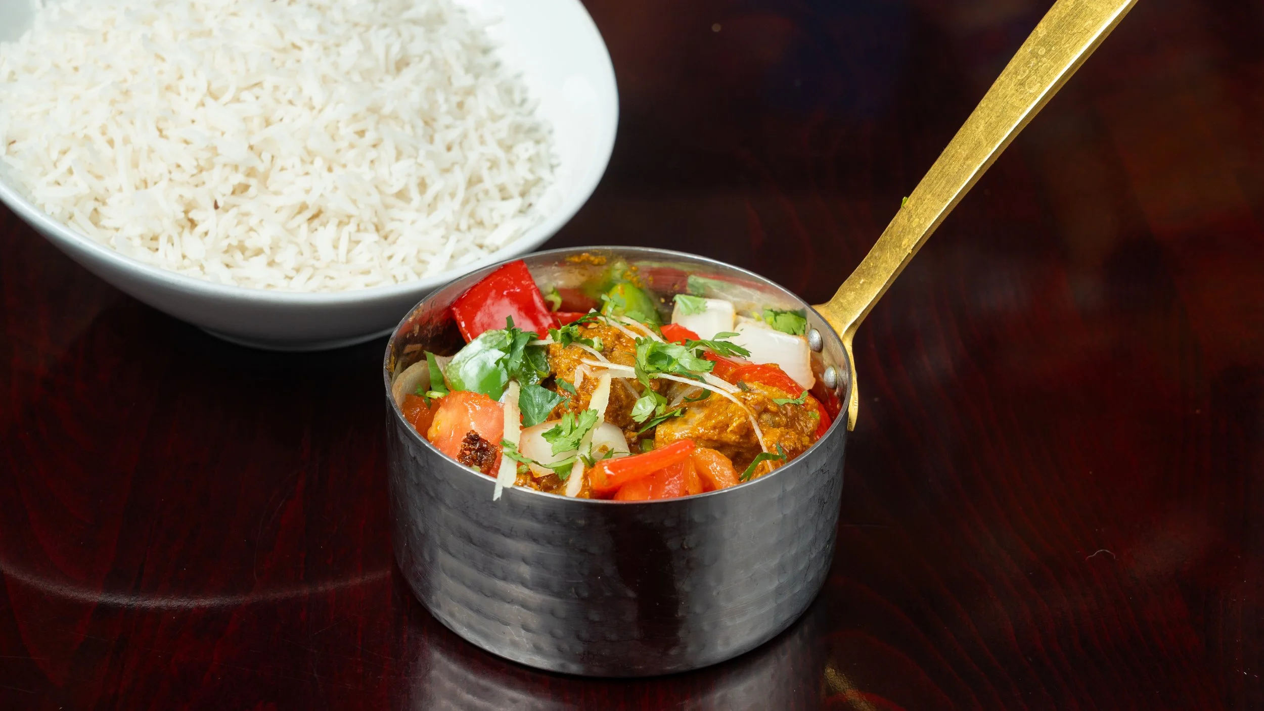 A bowl of white rice and a small metal pot of Indian vegetable curry garnished with cilantro on a dark wooden table.