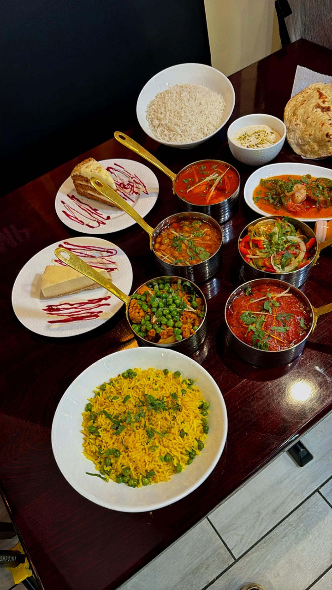 A table with Indian dishes including yellow rice with peas, various curry dishes in small bowls, naan bread, a slice of cake, a piece of cheese, and a bowl of rice.