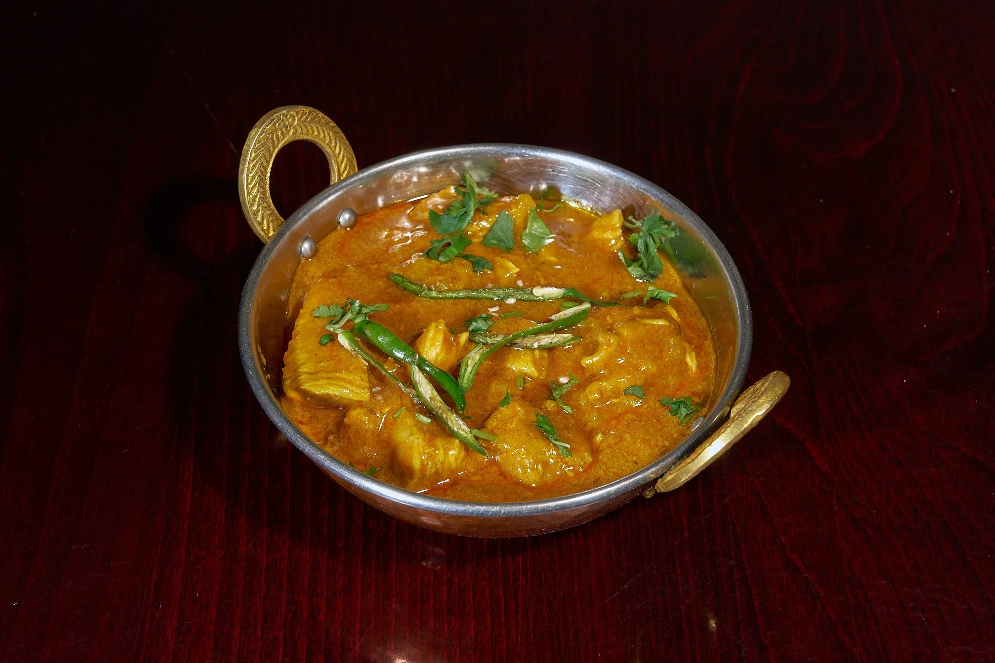 Indian curry dish with green chili peppers and cilantro in a silver bowl on a dark wooden table.