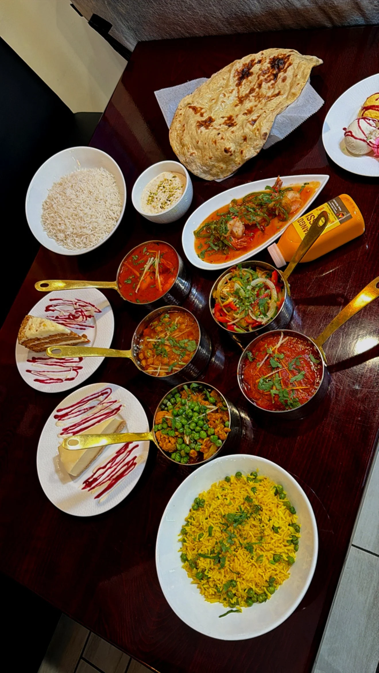 A variety of Indian dishes on a wooden table, including naan bread, basmati rice, yellow rice, curry dishes in bowls, a bowl of salad, and desserts, with condiments and utensils.