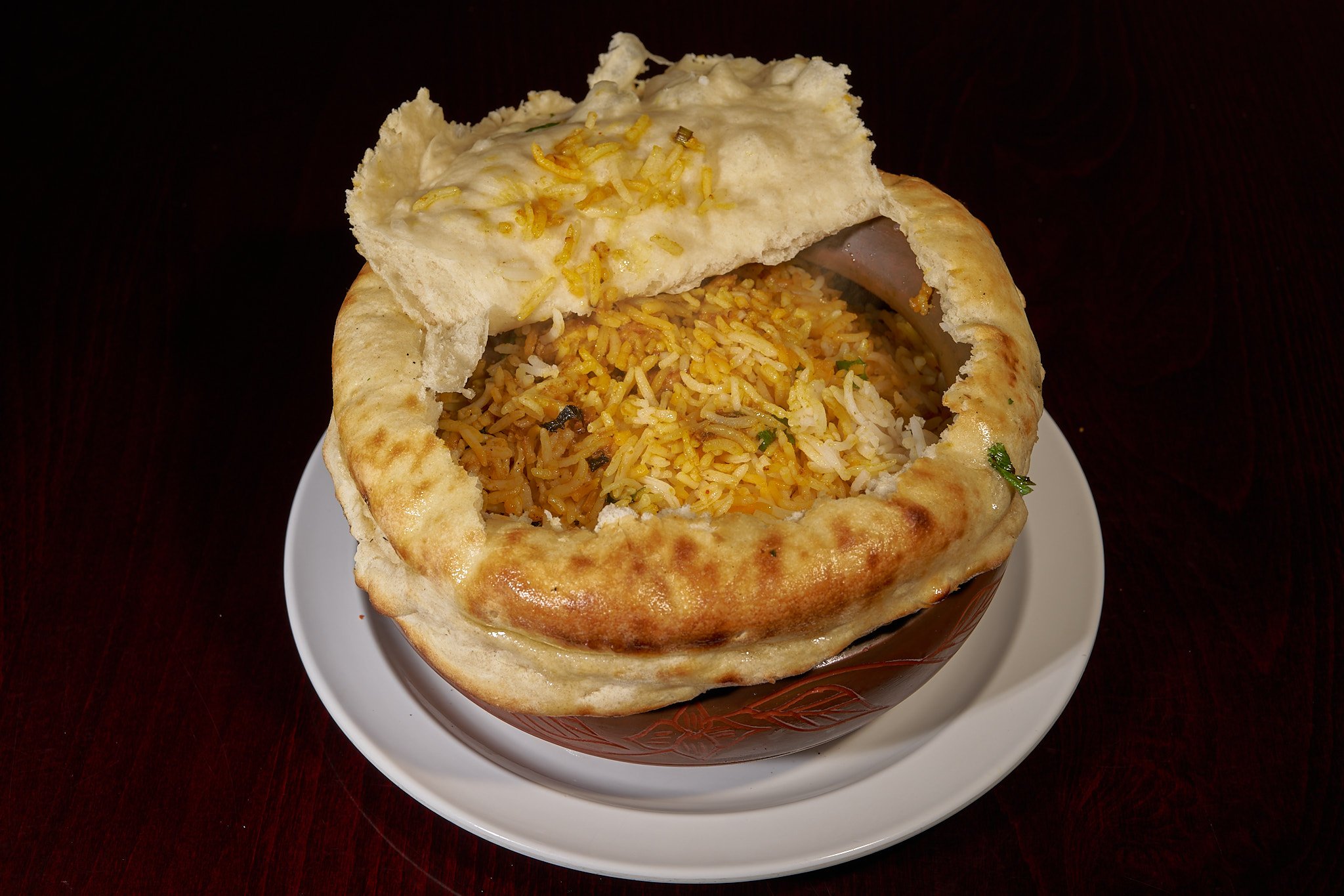 Bowl of Indian biryani rice with naan bread on top, served on a white plate on a dark wooden table.