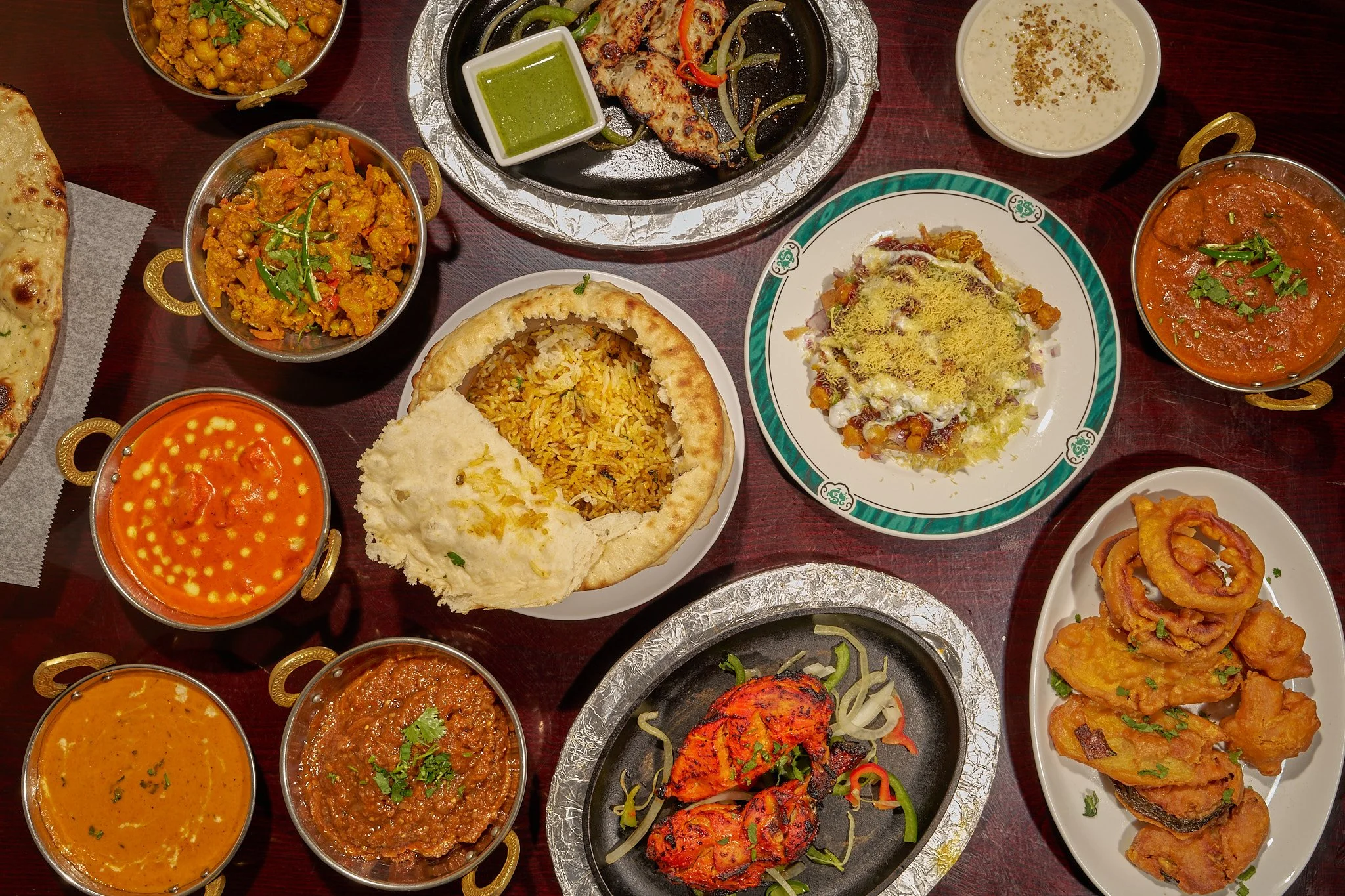An assortment of Indian dishes served on a table, including naan bread, rice, chicken tikka, various curries, and fried vegetables, with some sauces and condiments.
