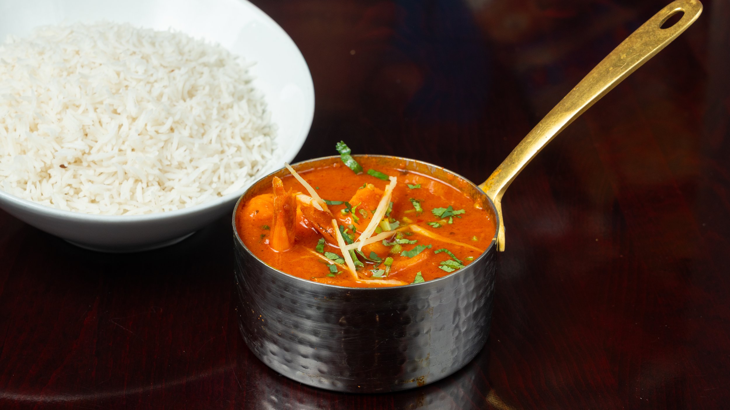A bowl of white rice next to a bowl of Indian curry garnished with cilantro and ginger on a dark wooden table.