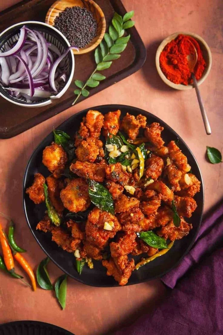 Fried chicken pieces garnished with garlic and curry leaves on a black plate, with chopped garlic and curry leaves, sliced red onions, red chili powder, dried red chili flakes, and fresh green chilies around it.