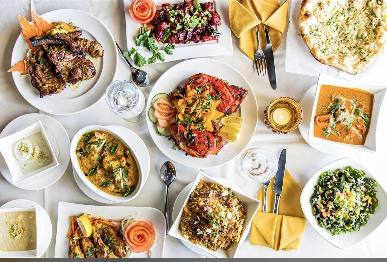 A table set with a variety of Indian dishes including grilled meats, curries, rice, naan, salad, and a dessert, with yellow napkins and glasses of water.
