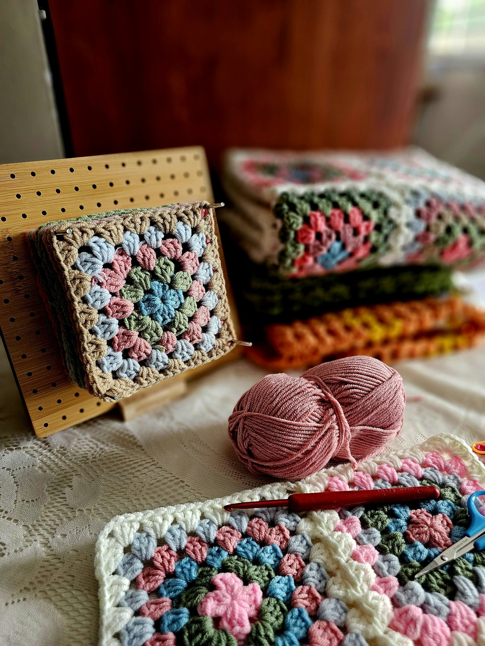 A table with crochet projects, yarn, scissors, and a crochet hook, with a stack of finished crochet squares and one in progress on a pegboard.