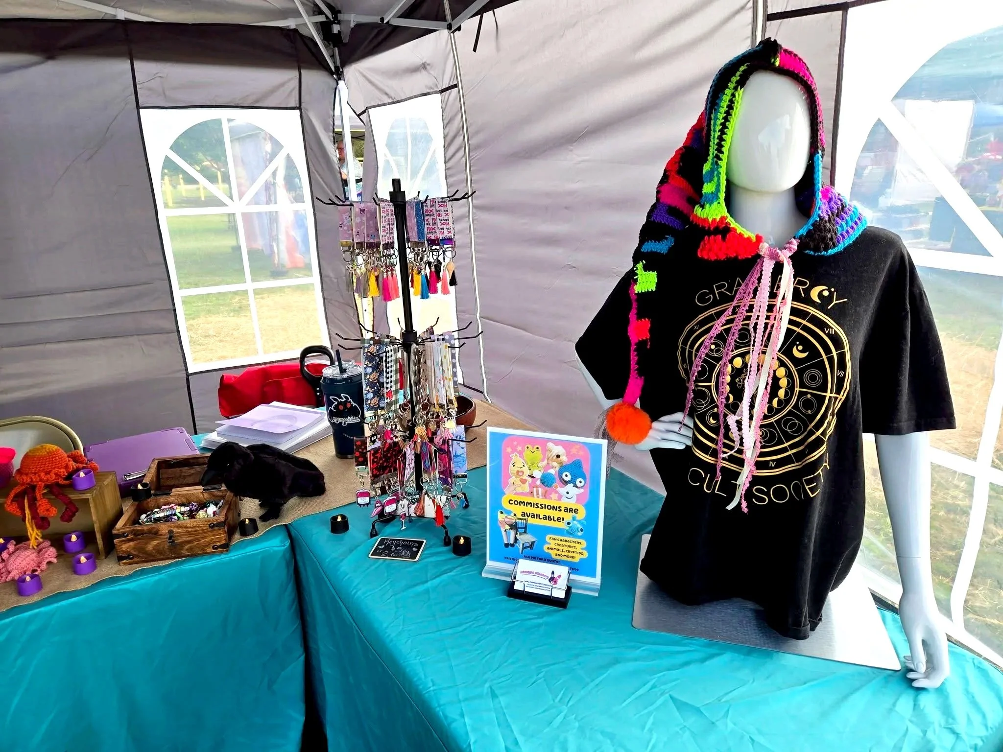 A booth at a fair displaying colorful handmade jewelry and accessories, with a mannequin wearing a multicolored hooded scarf and a signboard promoting fandom-themed merchandise and commissions.