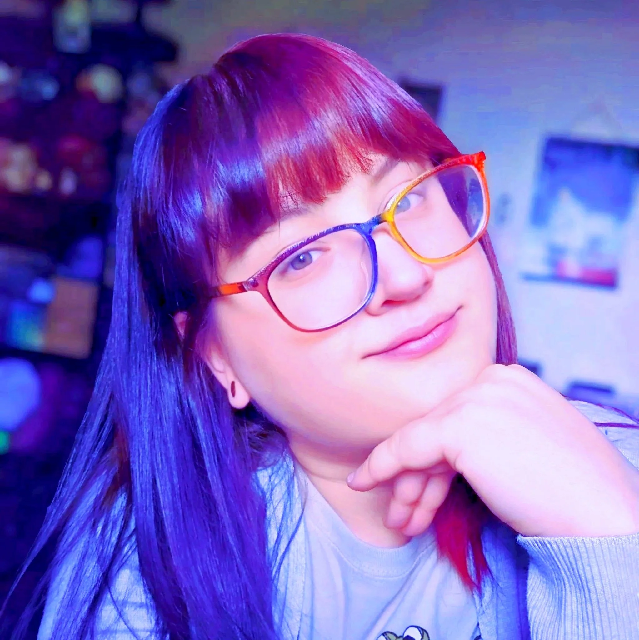 Close-up of a young woman with dyed purple and red hair, wearing colorful glasses and smiling softly, resting her chin on her hand.