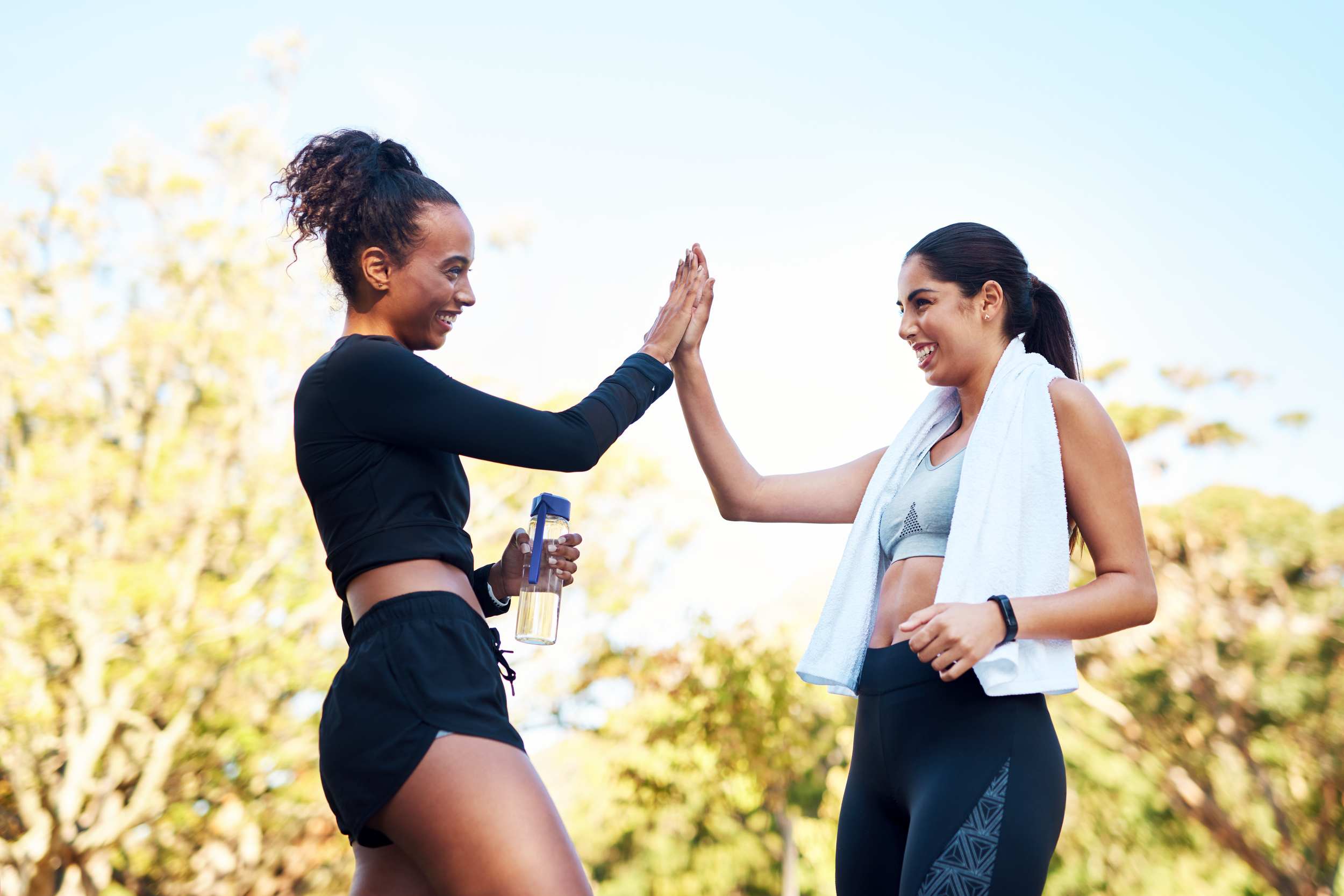 Two women smiling and giving each other a high five outdoors on a sunny day, one in athletic wear with a towel around her neck and the other in workout clothes holding a water bottle.
