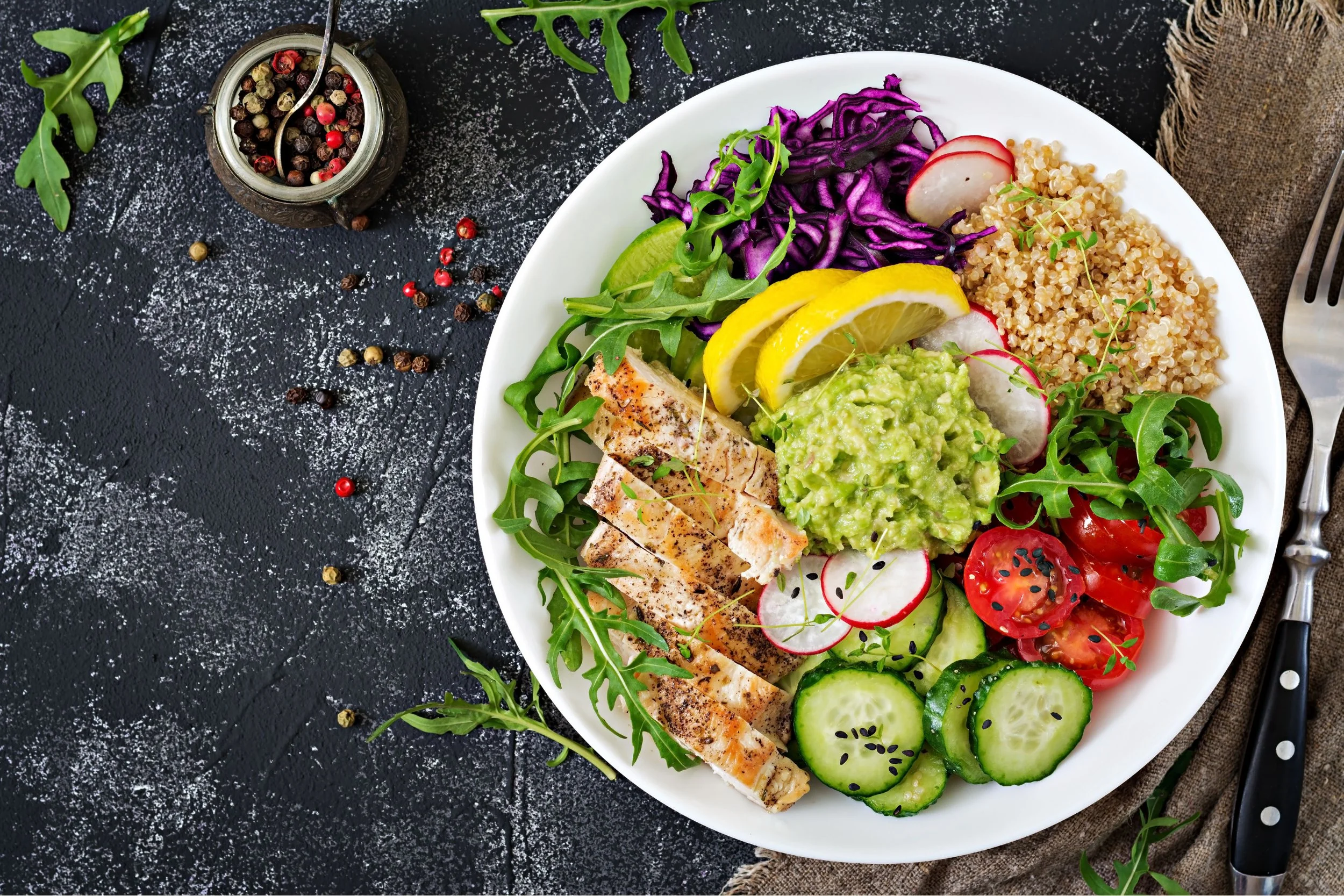 A white plate with a colorful salad including grilled chicken, cucumber, cherry tomatoes, radishes, shredded purple cabbage, lemon slices, and guacamole. There are green leaves and a rustic cloth to the right, and a fork and knife placed on the right side. The background surface is black with white powdered seasoning and scattered peppercorns, with some scattered on the surface.