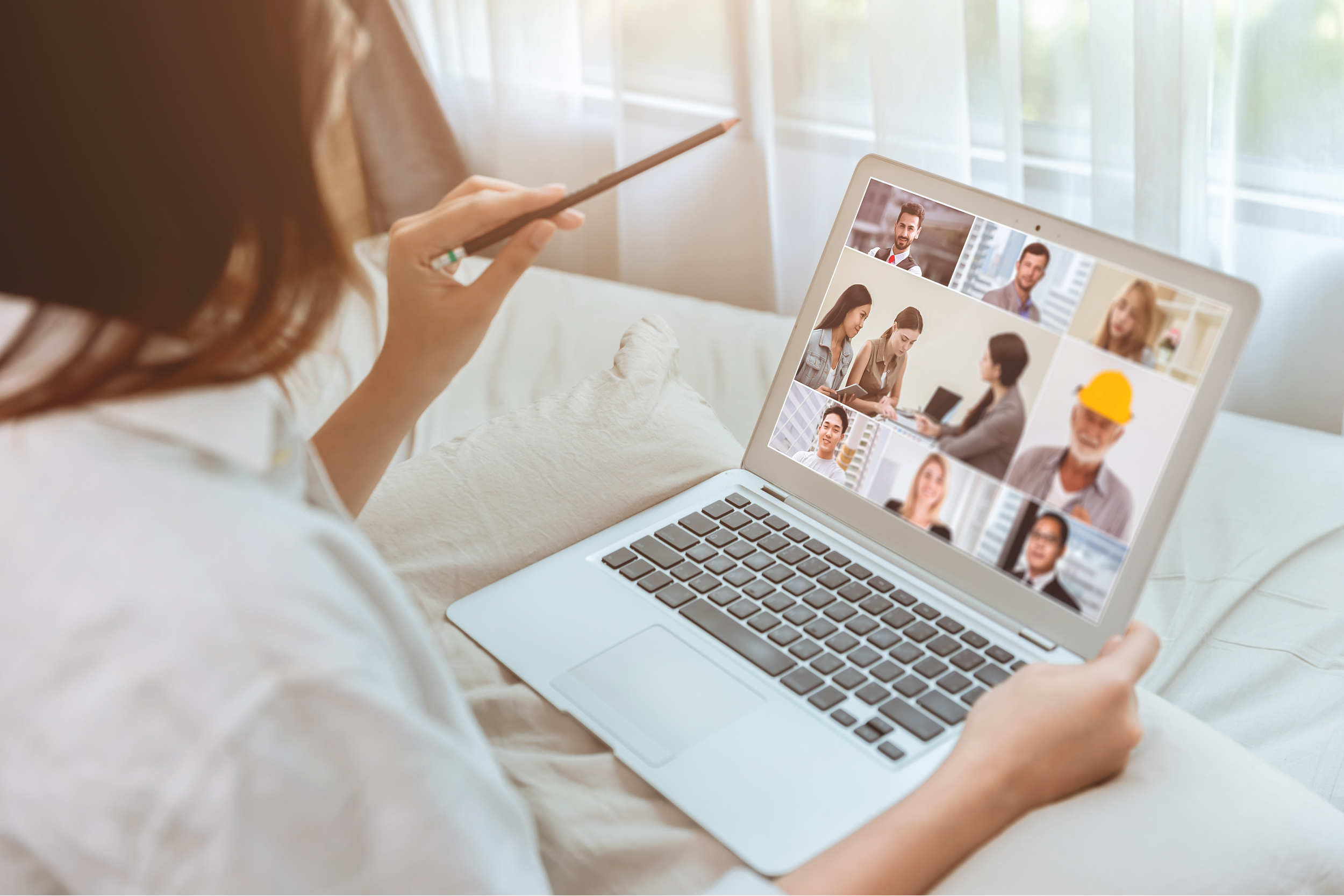 A woman sitting on a bed during daytime participating in a video call on her laptop, with several people shown in individual video windows on the screen.