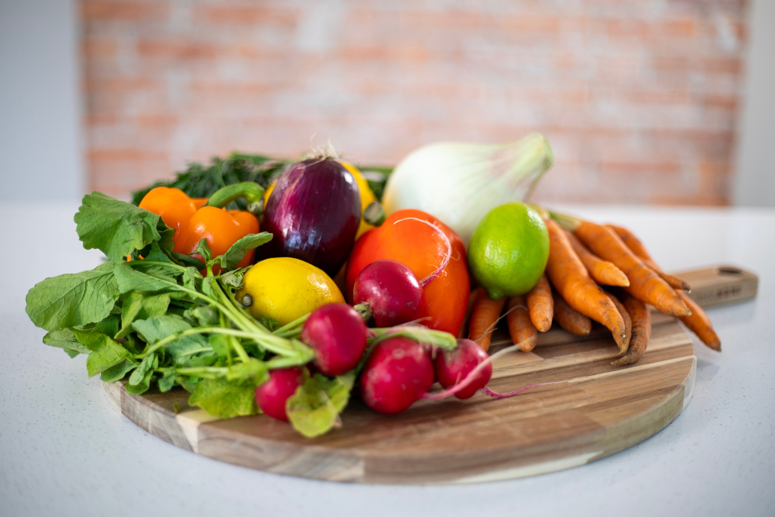 A wooden tray filled with fresh vegetables including radishes, carrots, a lime, an eggplant, a bell pepper, and an onion, with some greens and herbs, on a white surface against a background of a brick wall.
