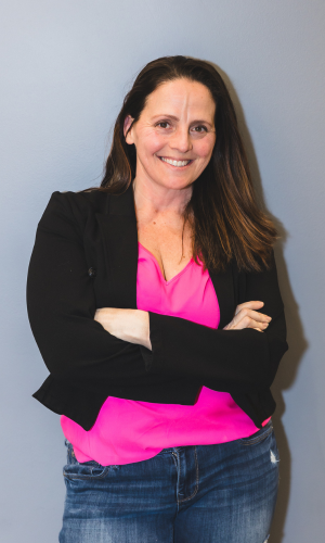 A woman with long brown hair, smiling and crossing her arms, standing against a light blue wall, wearing a black blazer, pink blouse, and blue jeans.