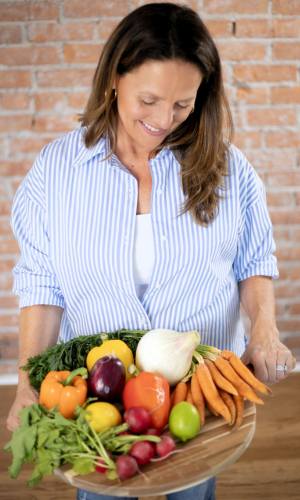 Woman smiling and holding a tray of fresh vegetables including carrots, radishes, apples, lemon, lettuce, and other produce