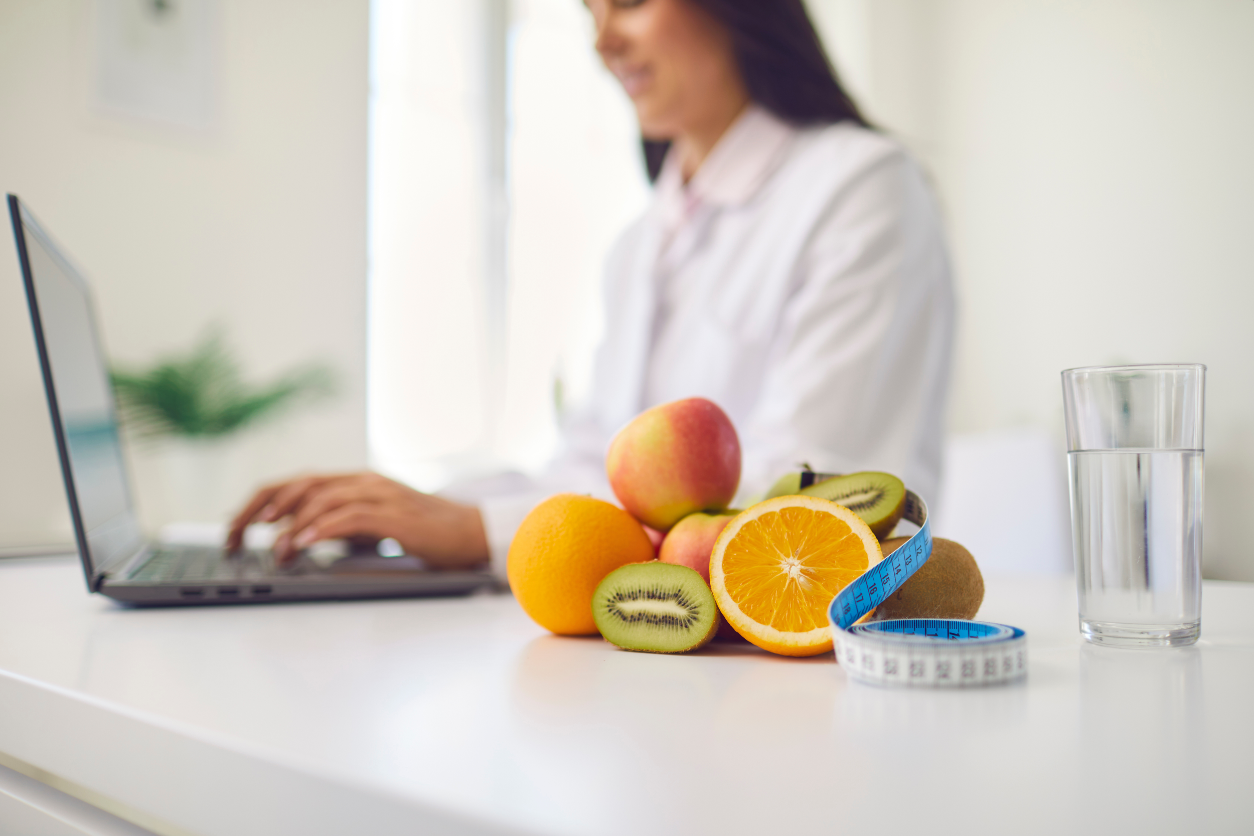 A white desk with a pile of fresh fruits including oranges, kiwis, and peaches, a blue measuring tape, and a glass of water, with a woman in the background working on a laptop, blurred image.