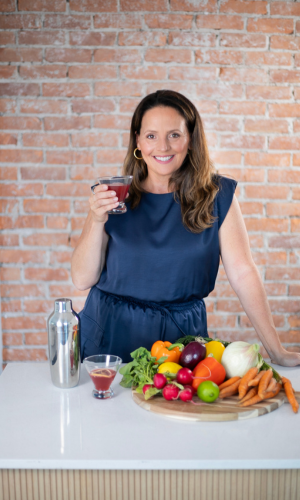 A woman in a blue dress holding a glass of red drink, standing behind a table with various vegetables and a cocktail shaker, against a brick wall background.