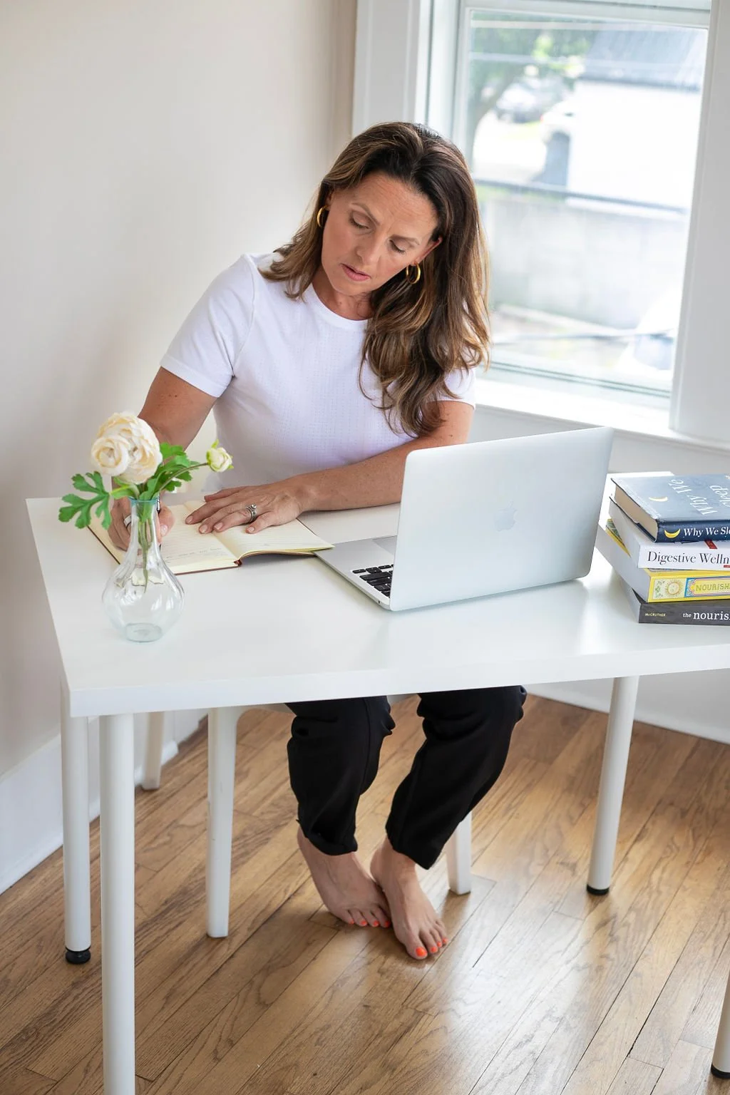 A woman sitting at a white desk, working with a laptop and reading a book, in a bright room with a large window, a vase with white flowers, and a stack of books.