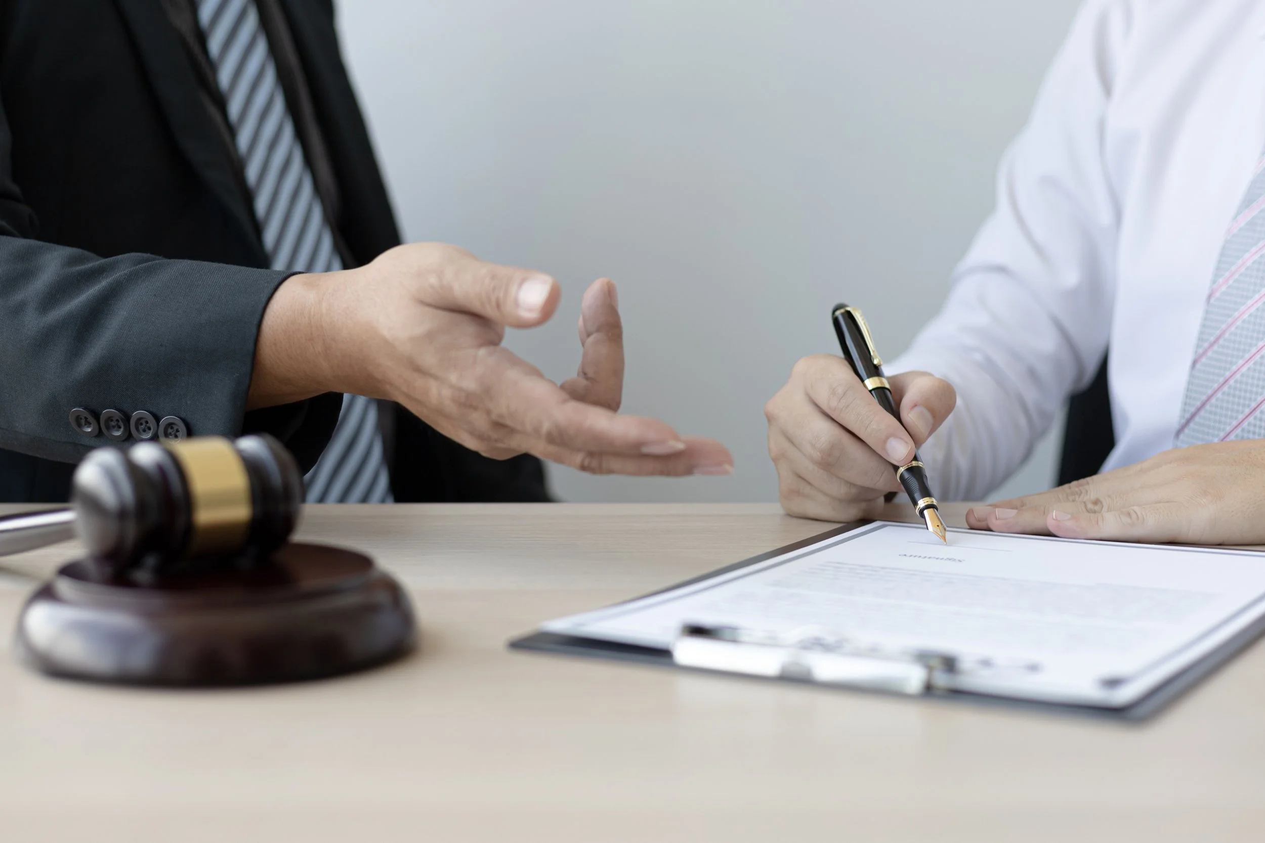 Close-up of two individuals in a professional setting, one wearing a dark suit and striped tie, the other in a white shirt and light-colored tie, engaged in a handshake. A gavel, documents, and a pen are on the table, indicating a legal or official meeting.