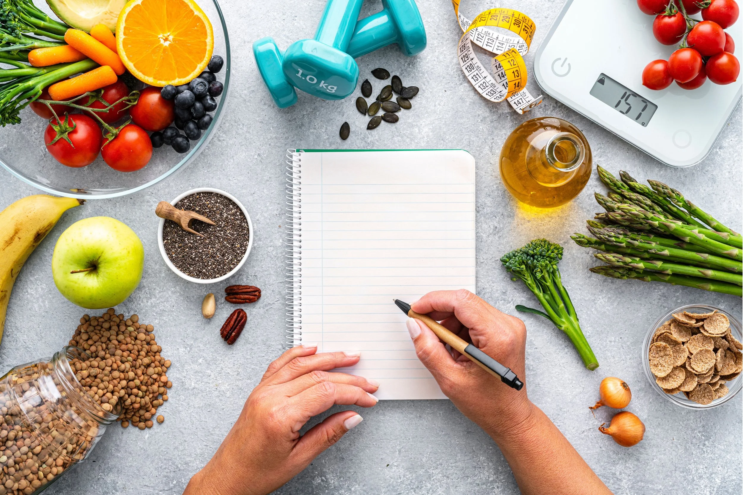A person is writing in a notebook surrounded by healthy foods and workout equipment, including a bowl of assorted vegetables and fruits, dumbbells, measuring tape, a digital scale with cherry tomatoes, a jar of oil, asparagus, onions, dried cereal, and seeds.