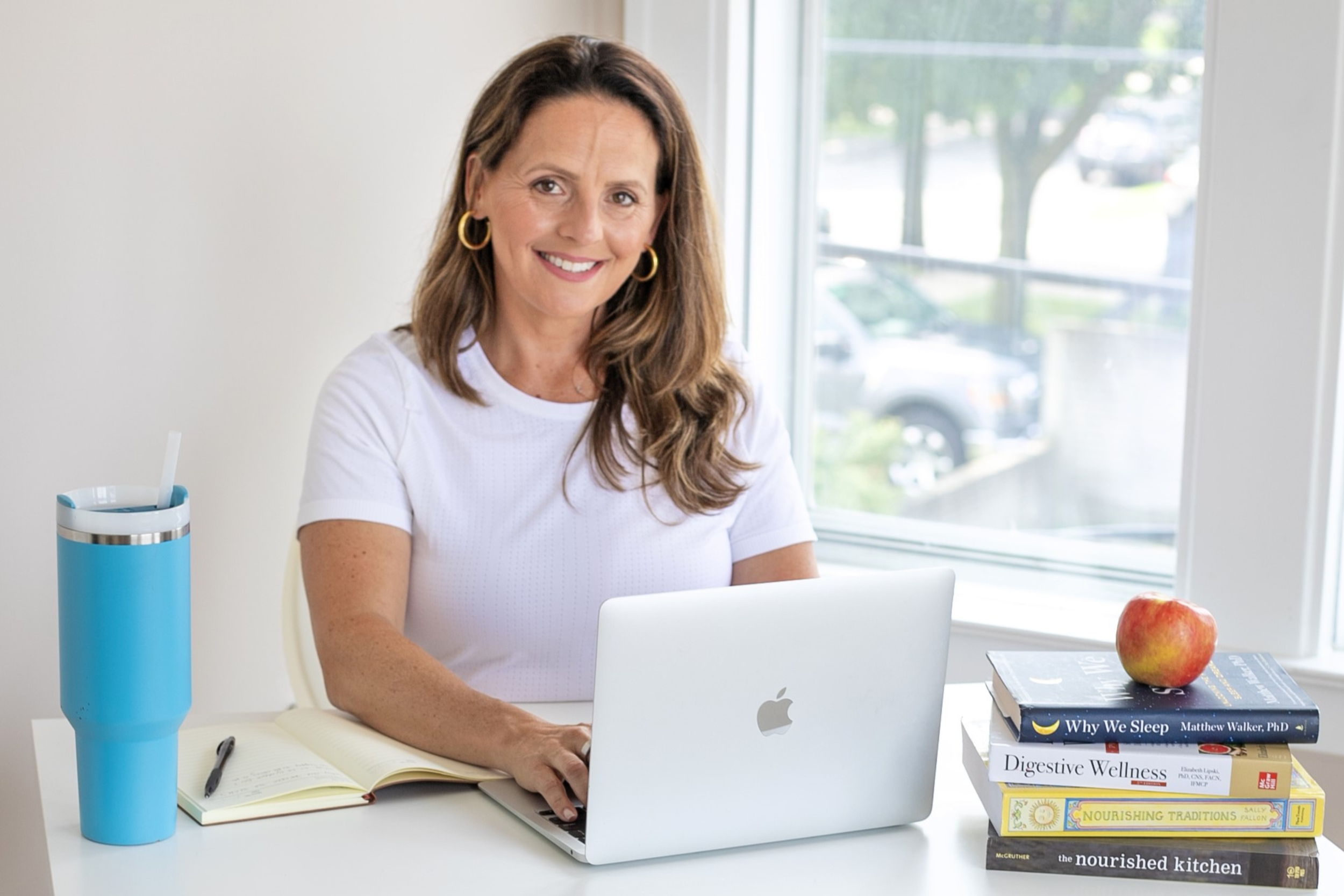 Woman with brown hair and gold hoop earrings working on a MacBook at a white desk with a stack of books, a blue water bottle, and an apple, near a window.