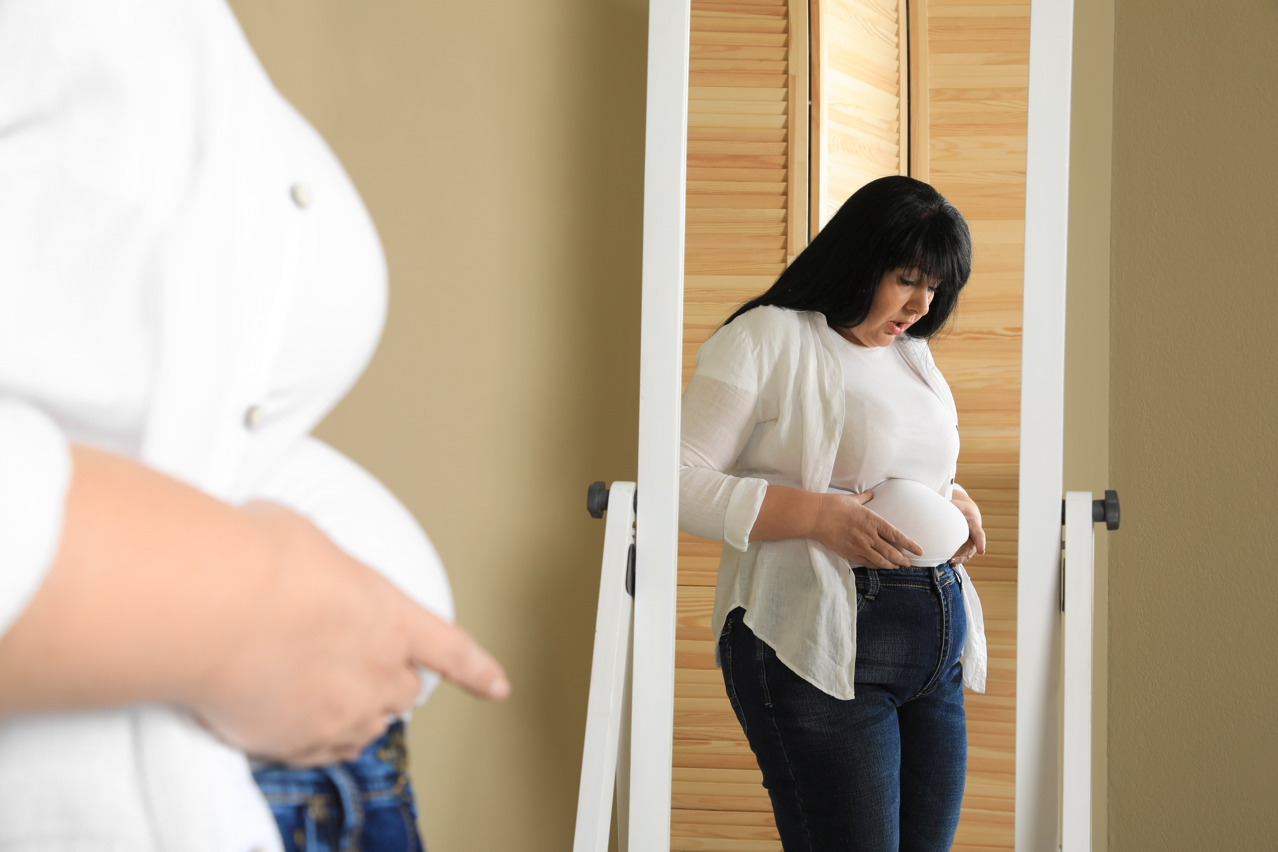 A woman looking down at her pregnant belly in front of a mirror in a bedroom, with a person in a white shirt partially visible in the foreground.