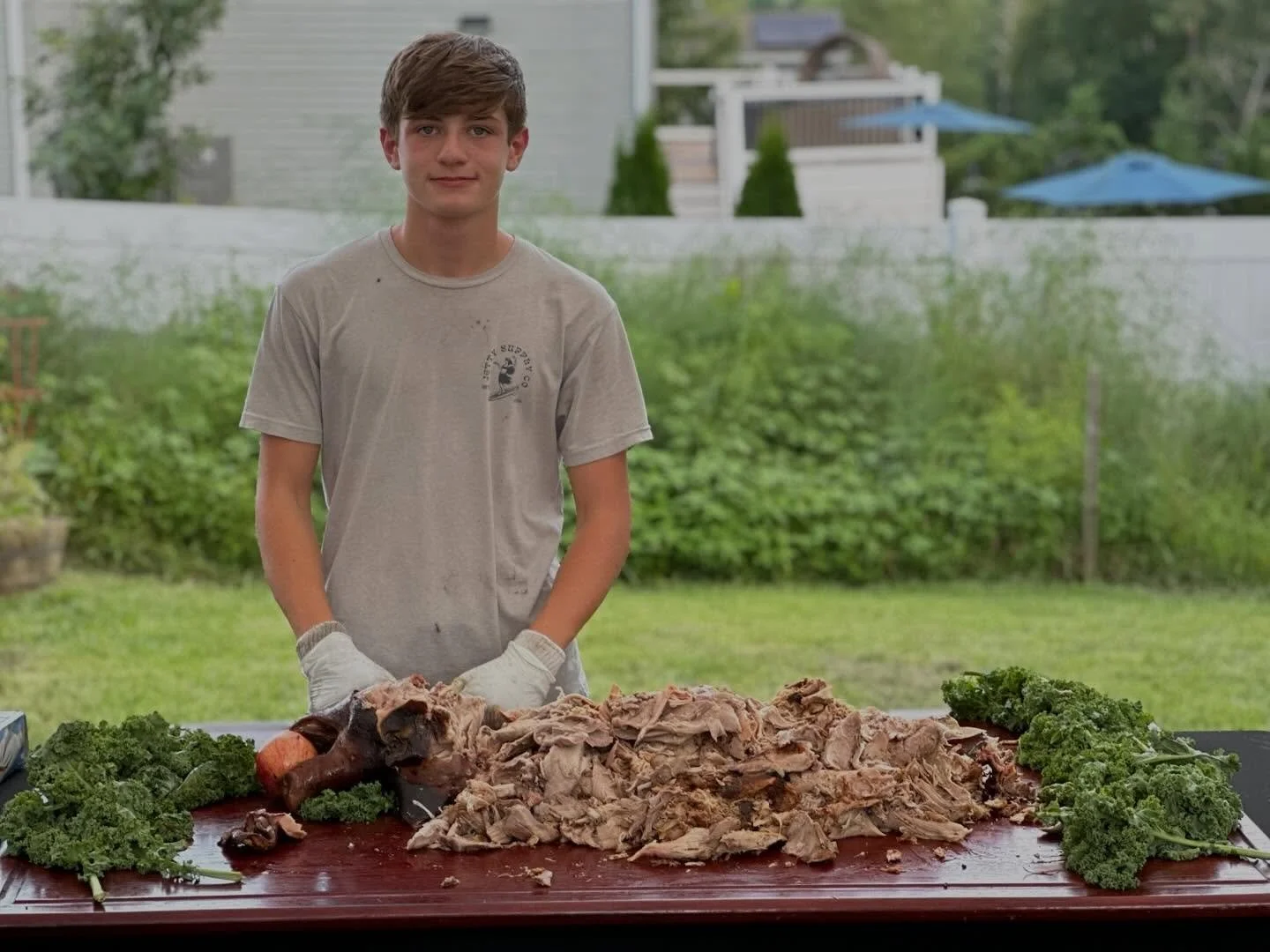 A teenage boy wearing a gray t-shirt and gloves stands behind a large cutting board with cooked, shredded meat and parsley in a backyard.