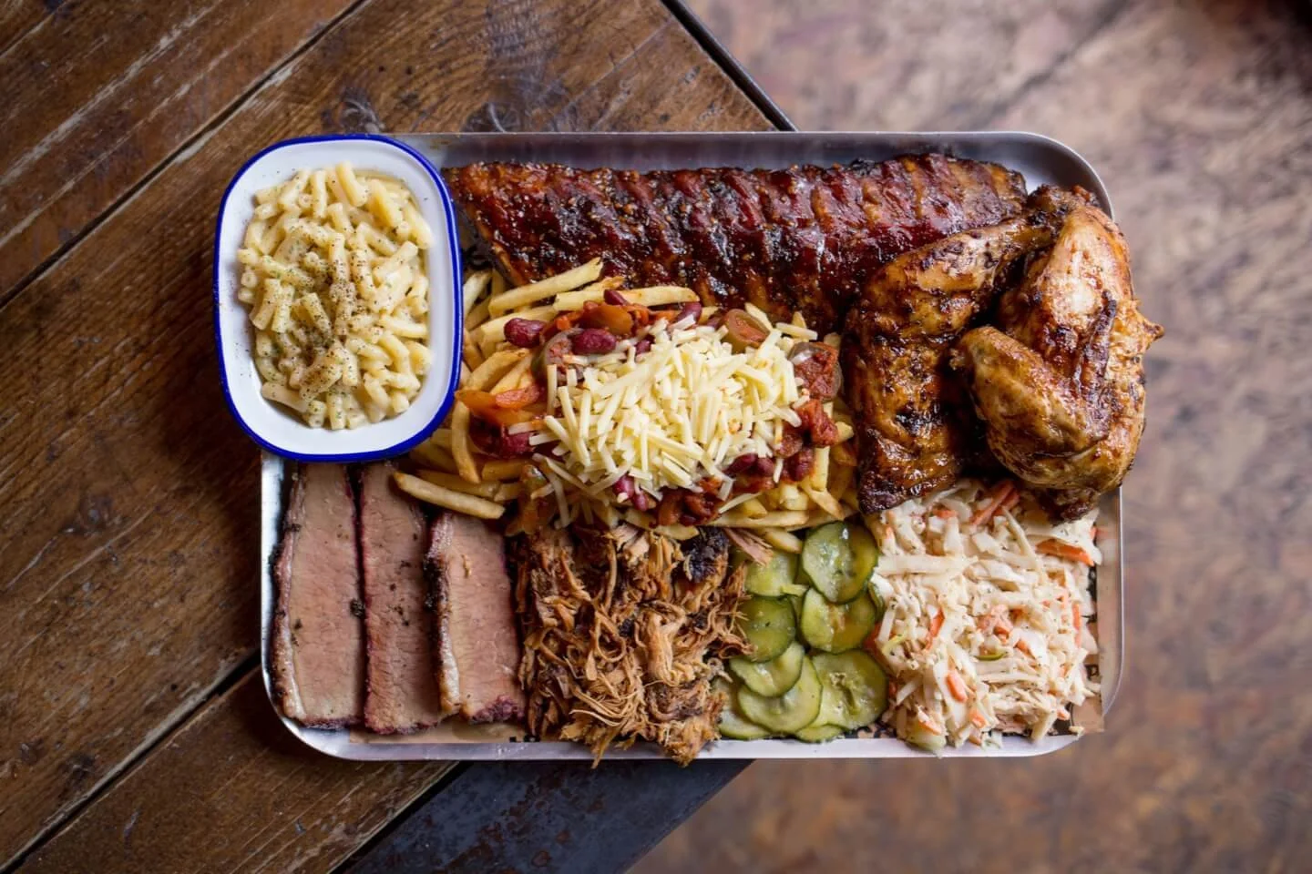 A tray with various barbecue foods including ribs, chicken, brisket, beans, coleslaw, pickles, pasta salad, and shredded cheese.